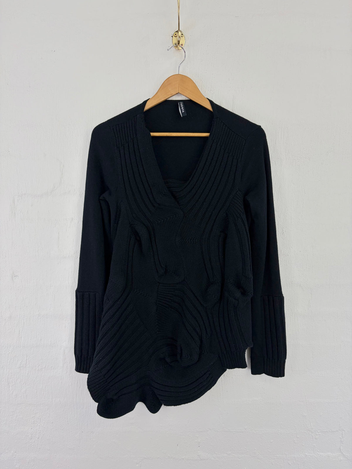 A black long-sleeve knit cardigan sweater with textured swirl patterns, hanging on a wooden hanger against a white painted brick wall.