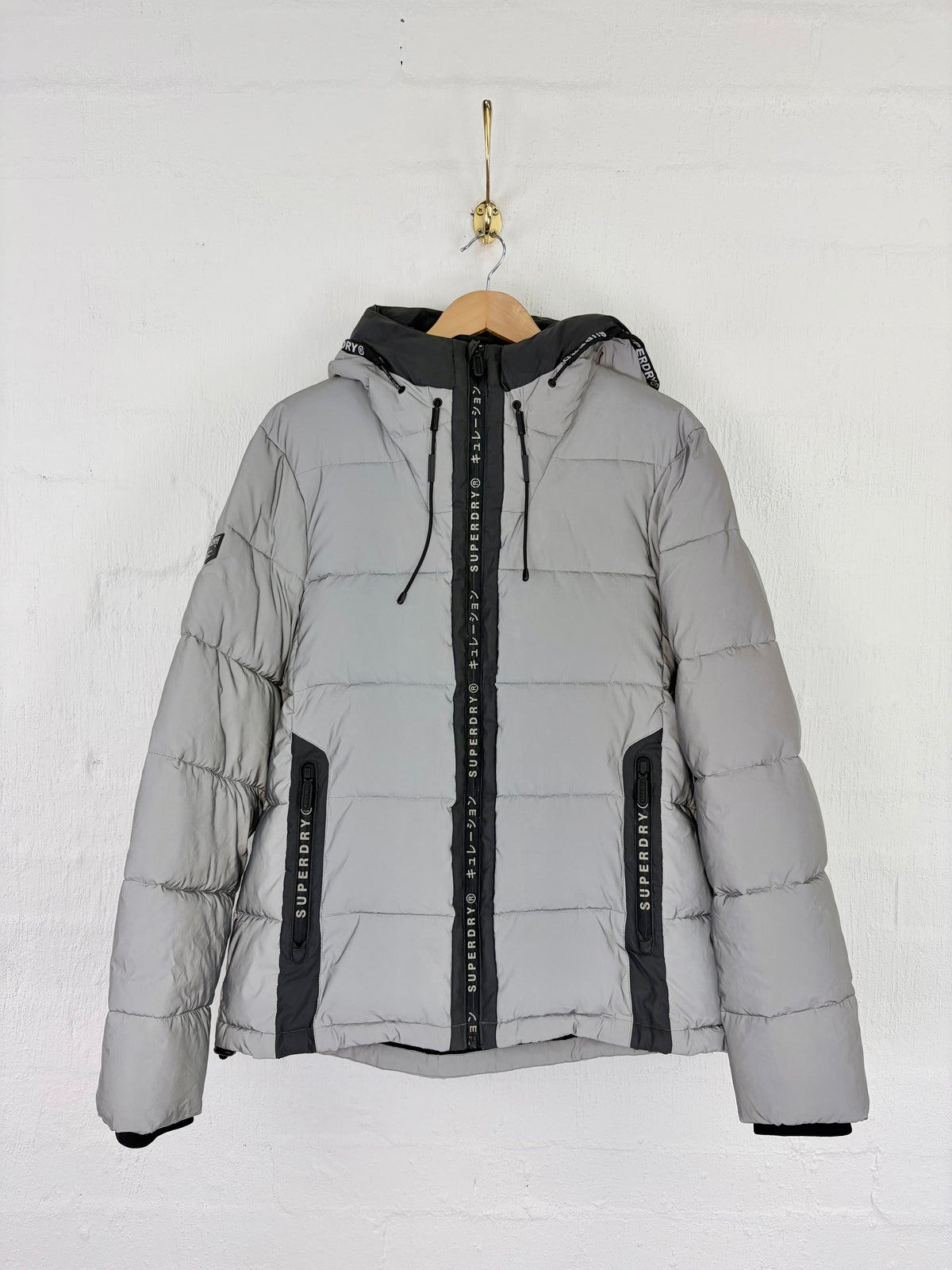 A light gray hooded puffer jacket with black trim and a black front zipper, hanging on a wooden hanger against a white brick wall.