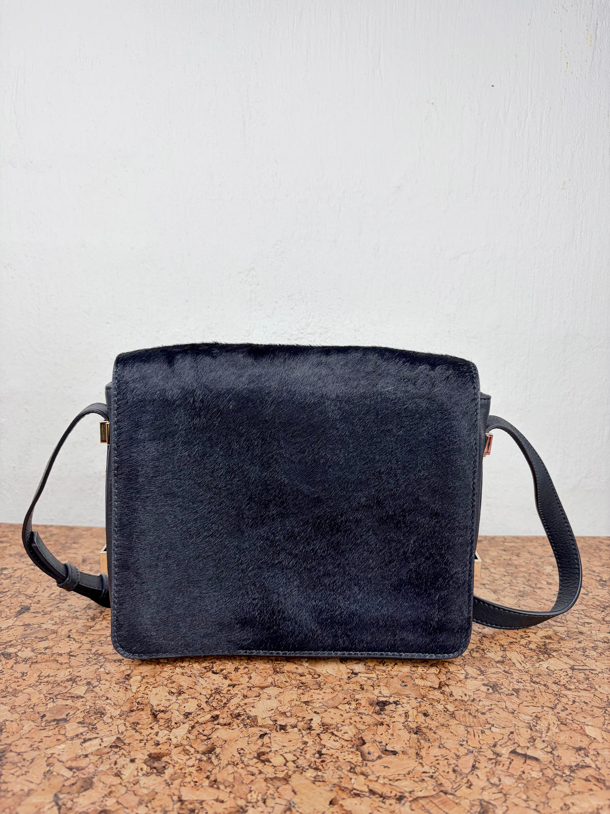 A black faux fur shoulder bag with a black strap, resting on a cork surface against a white wall.