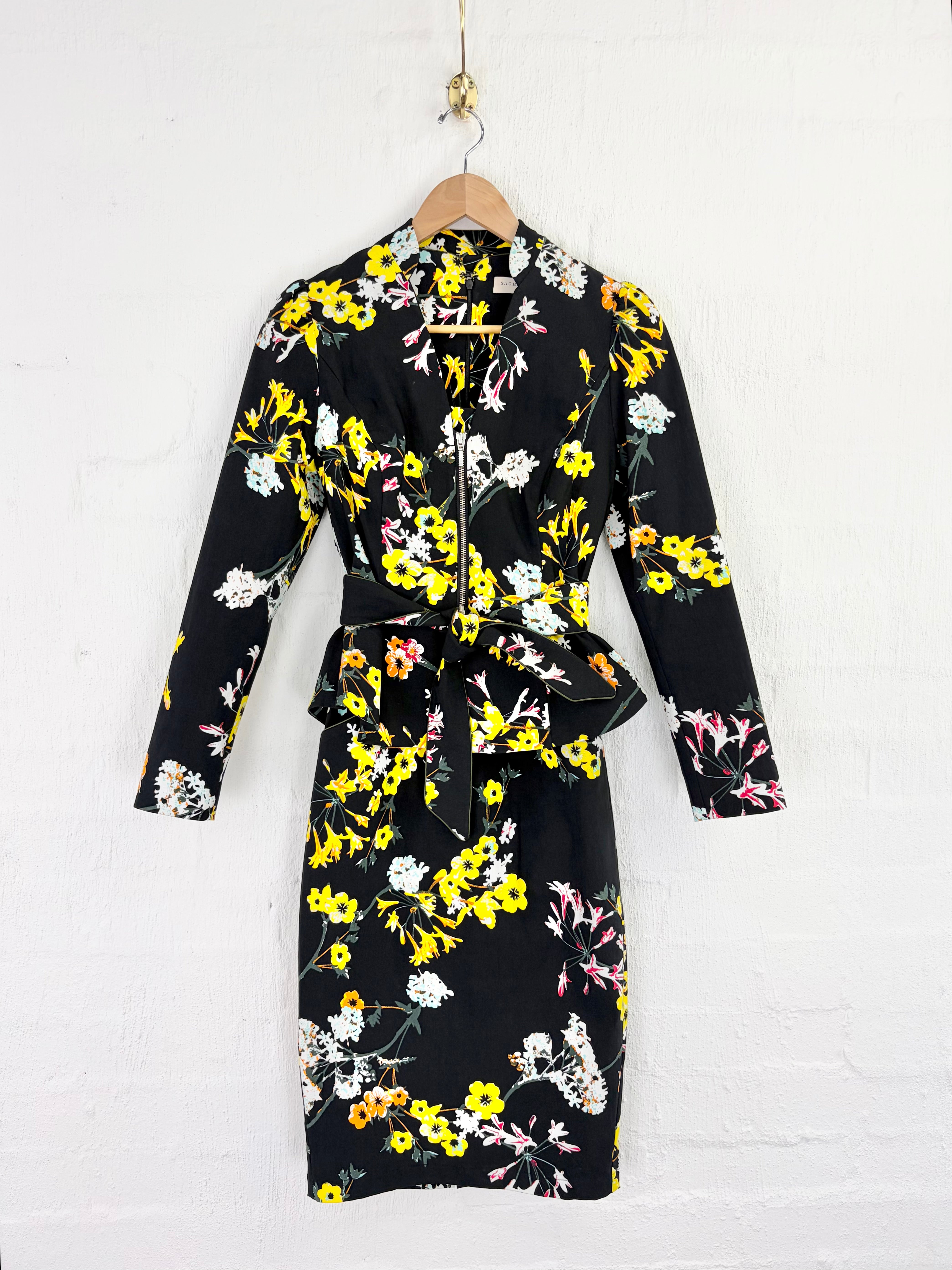 A black long-sleeve wrap dress with a vibrant yellow, white, and pink floral print, hanging on a wooden hanger against a white painted brick wall.