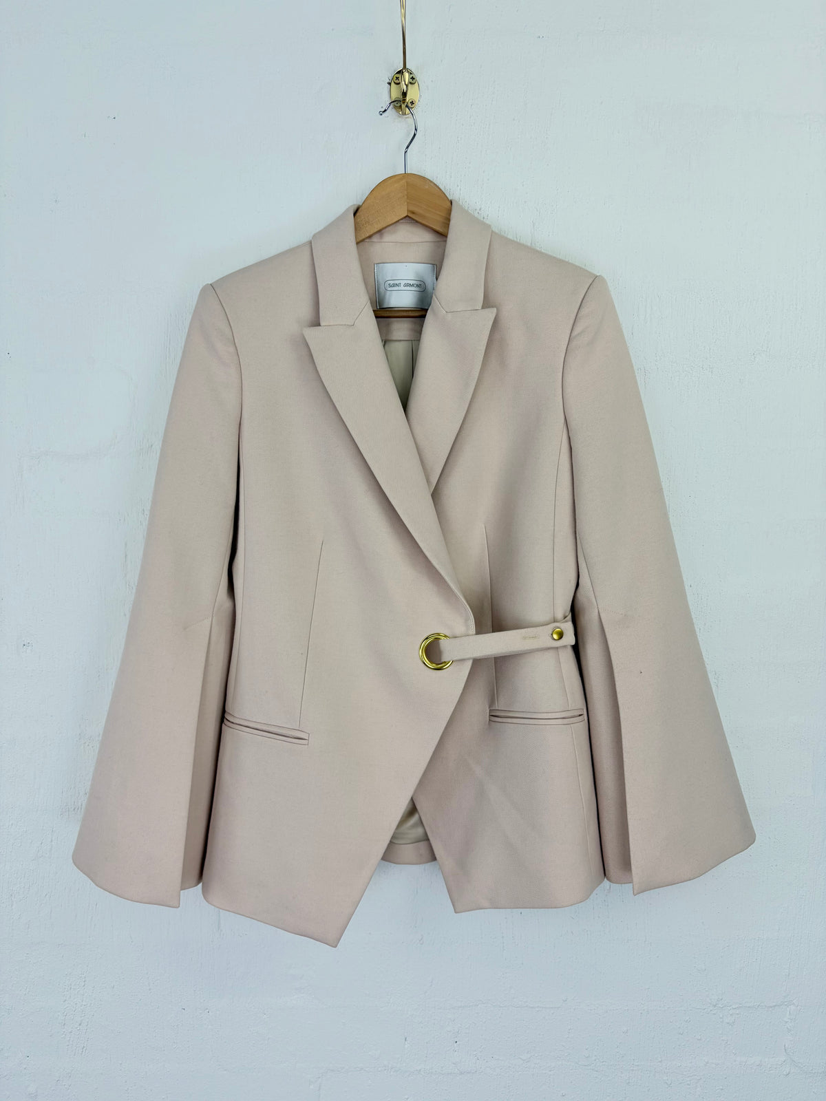 A light beige wrap-style blazer with a matching belt and gold-tone ring closure, hanging on a wooden hanger against a white painted brick wall.