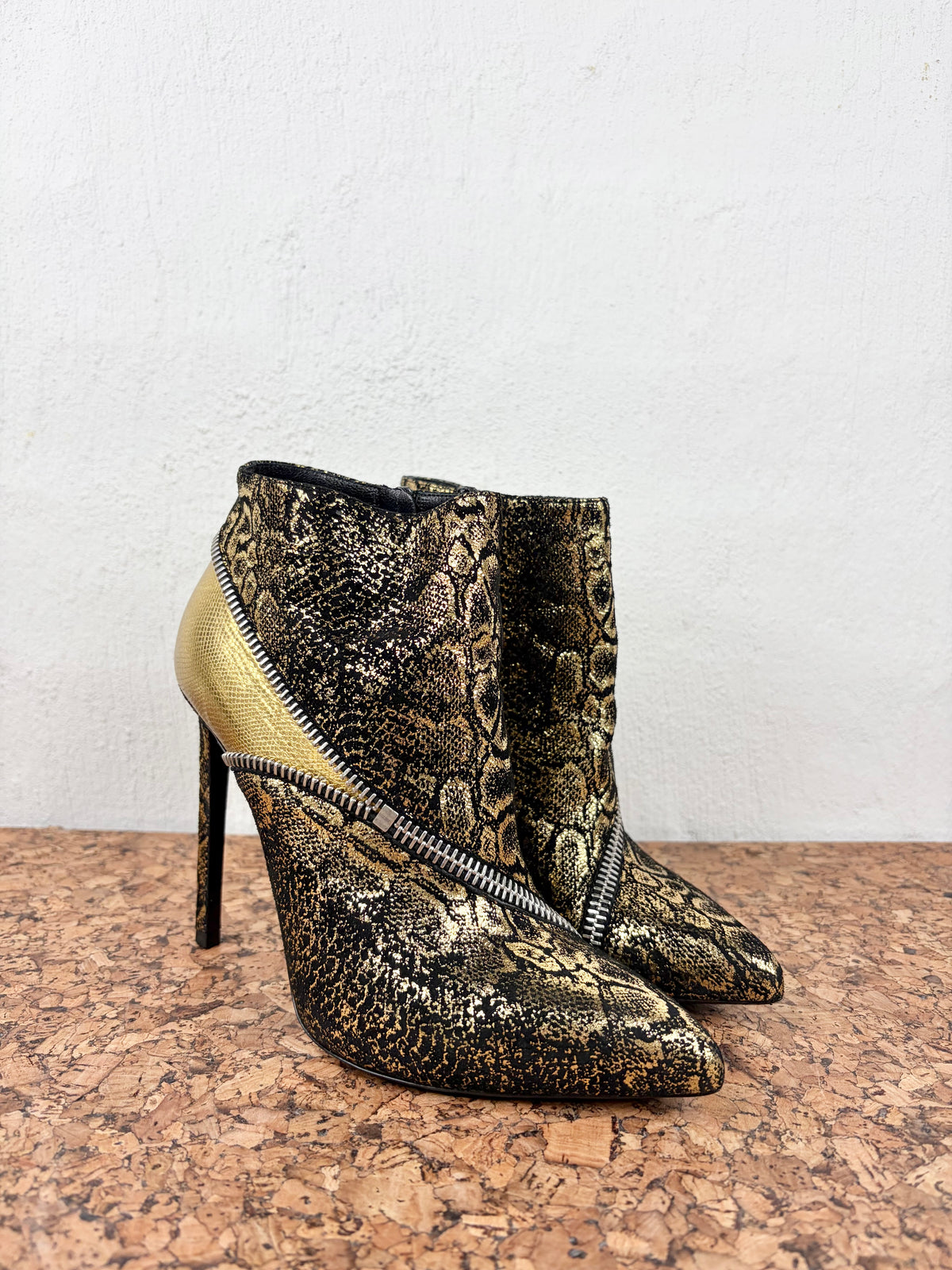 A pair of pointed-toe high-heeled ankle boots featuring a metallic gold and black snakeskin pattern, with a contrasting smooth gold side panel and a silver zipper detail across the front, set on a cork surface against a white wall.