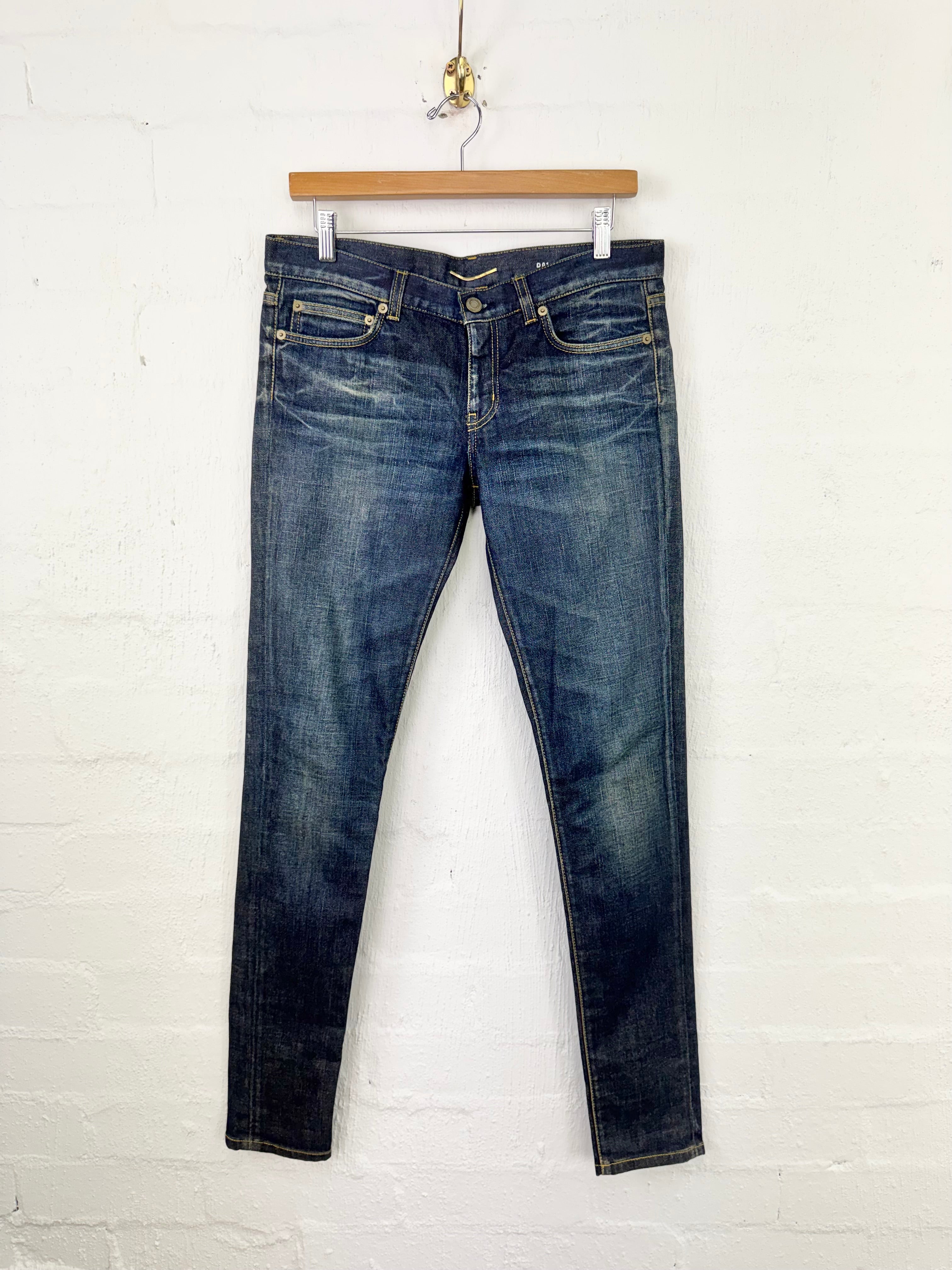 A pair of dark blue slim-fit jeans with light wash fading on the thighs, hanging from a wooden hanger against a white painted brick wall.