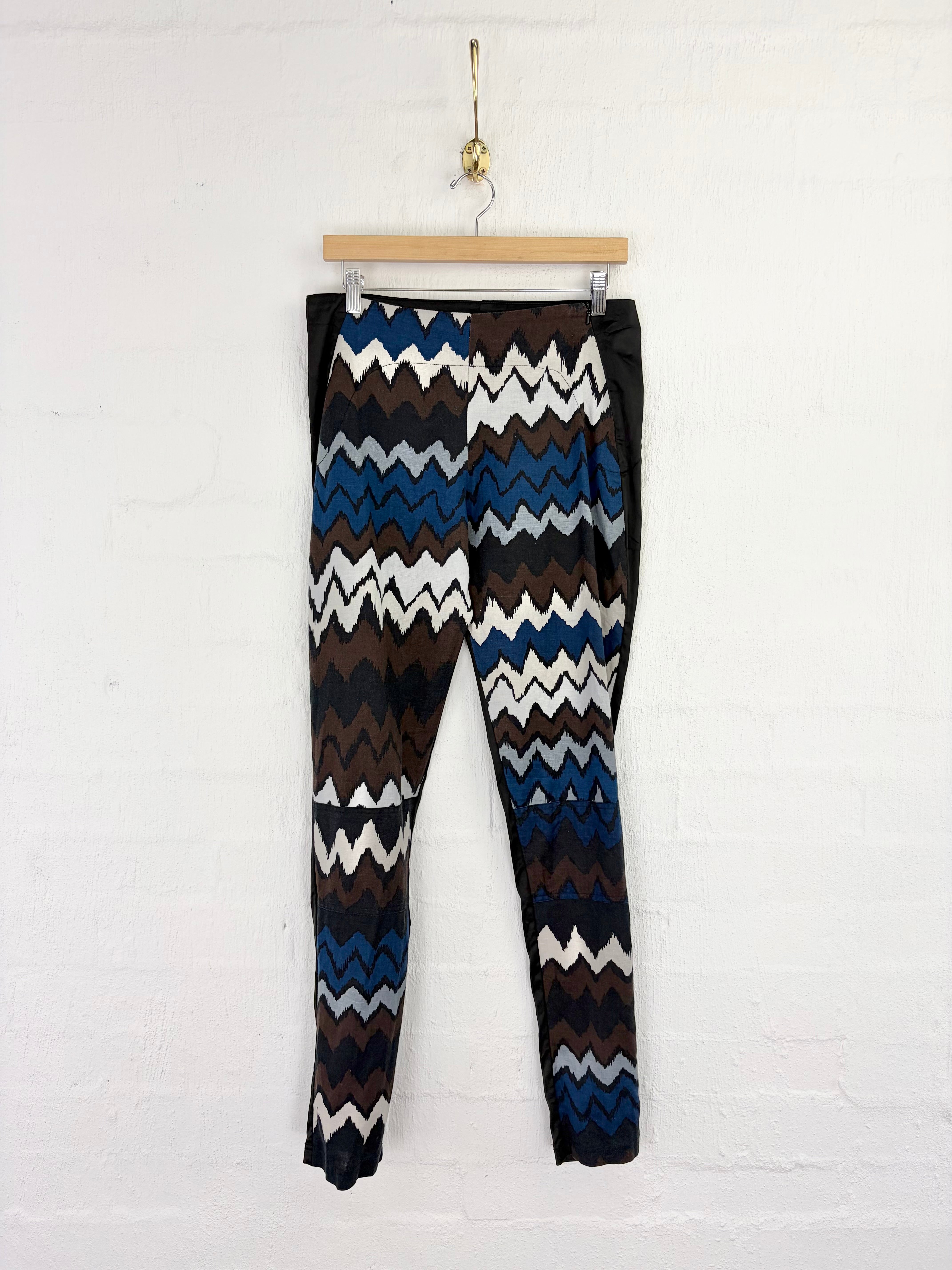 Patterned knit pants hanging from a wooden hanger against a white painted brick wall. The pants feature a bold chevron pattern in blue, brown, white, and black, with solid black side panels.