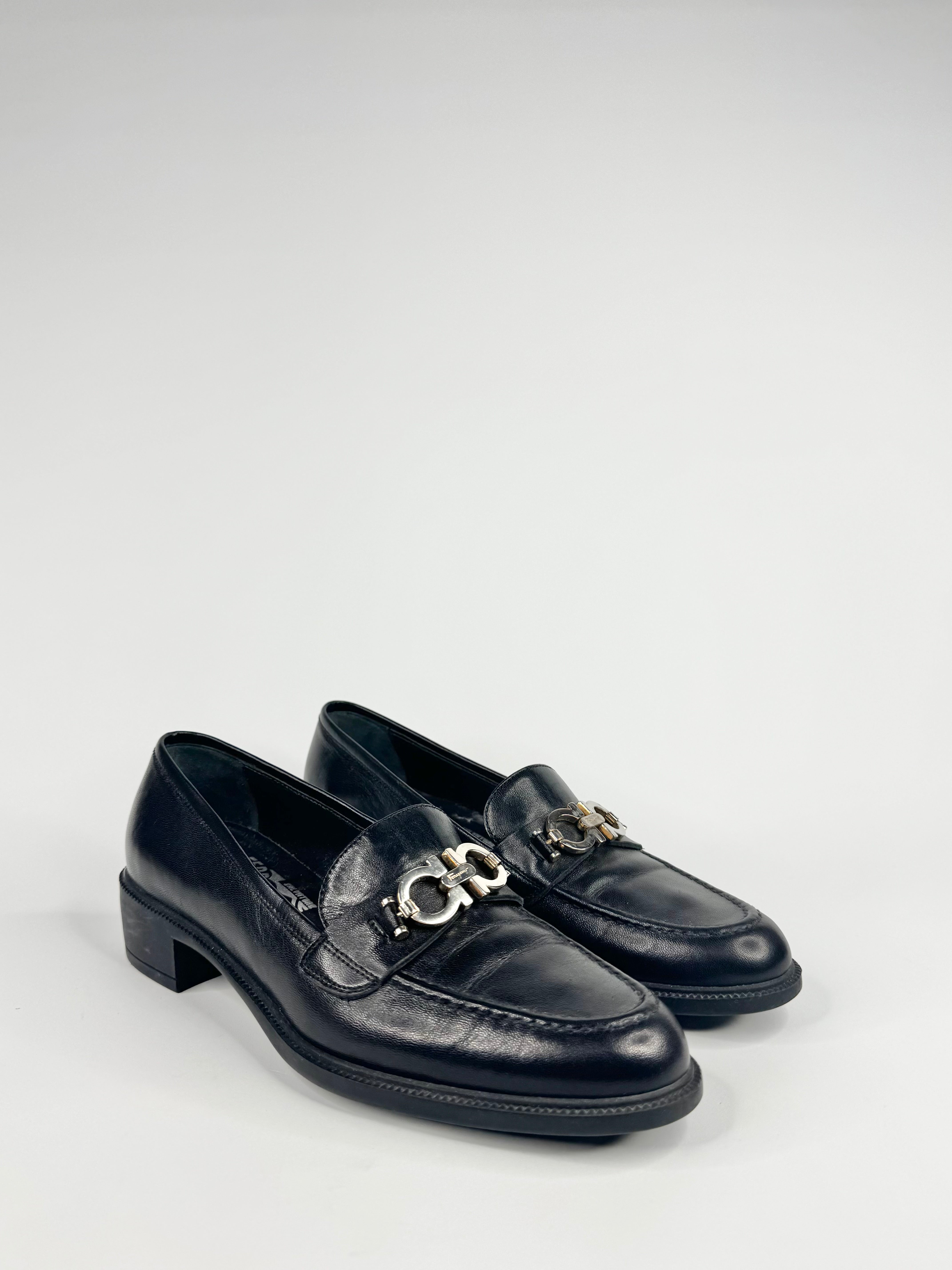 Pair of black loafers with gold chain detail on a light gray background