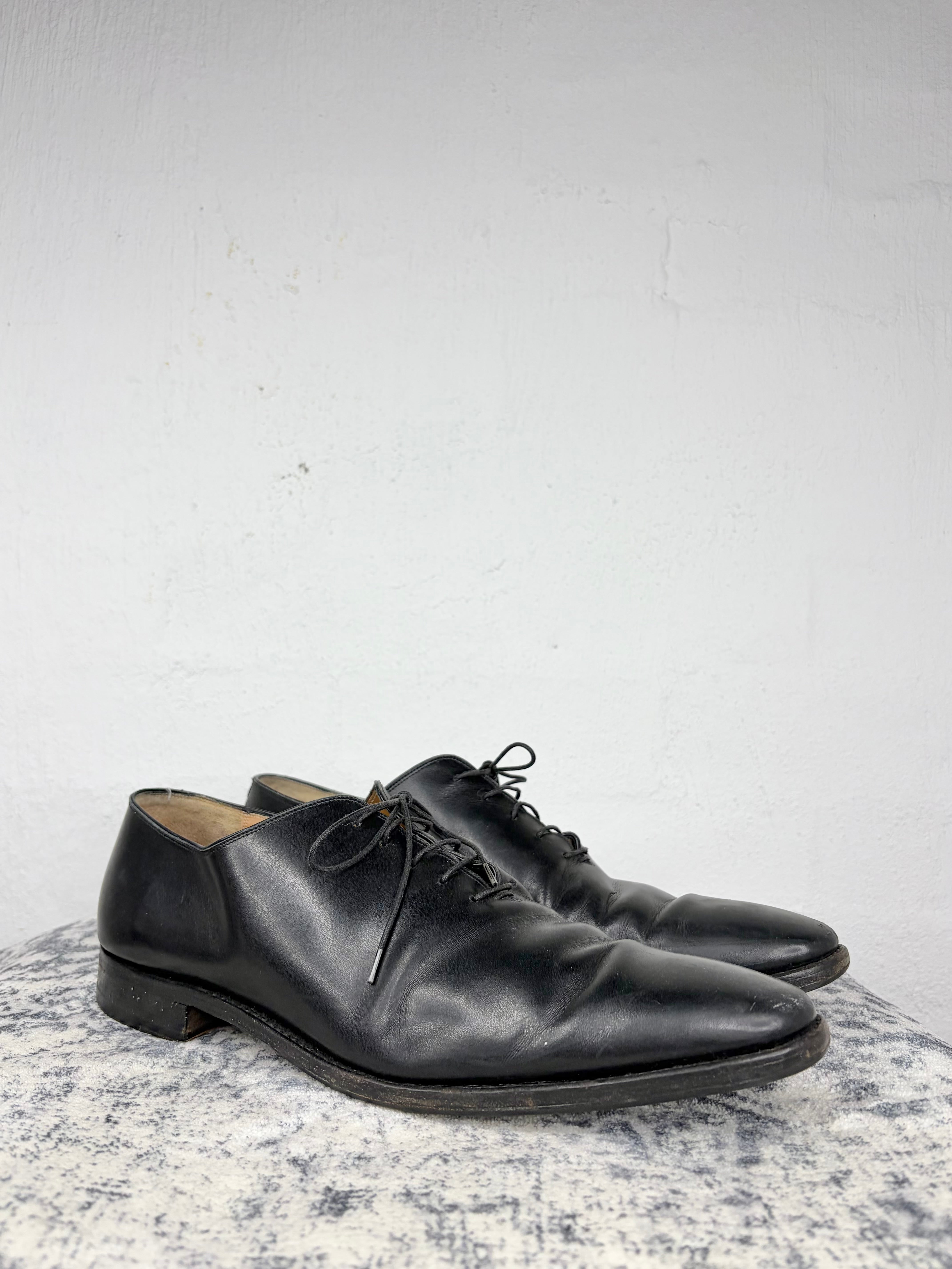 A pair of black leather oxford shoes with closed lacing, displayed on a textured gray stone surface against a white wall.