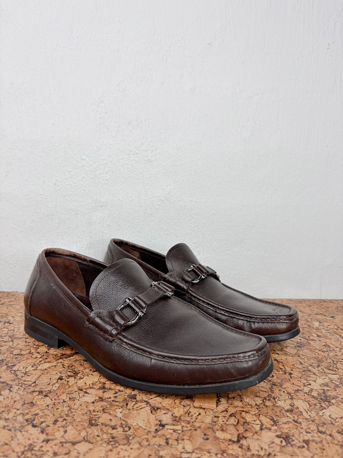 A pair of dark brown leather penny loafers with double strap buckles, displayed on a cork surface against a light gray wall.
