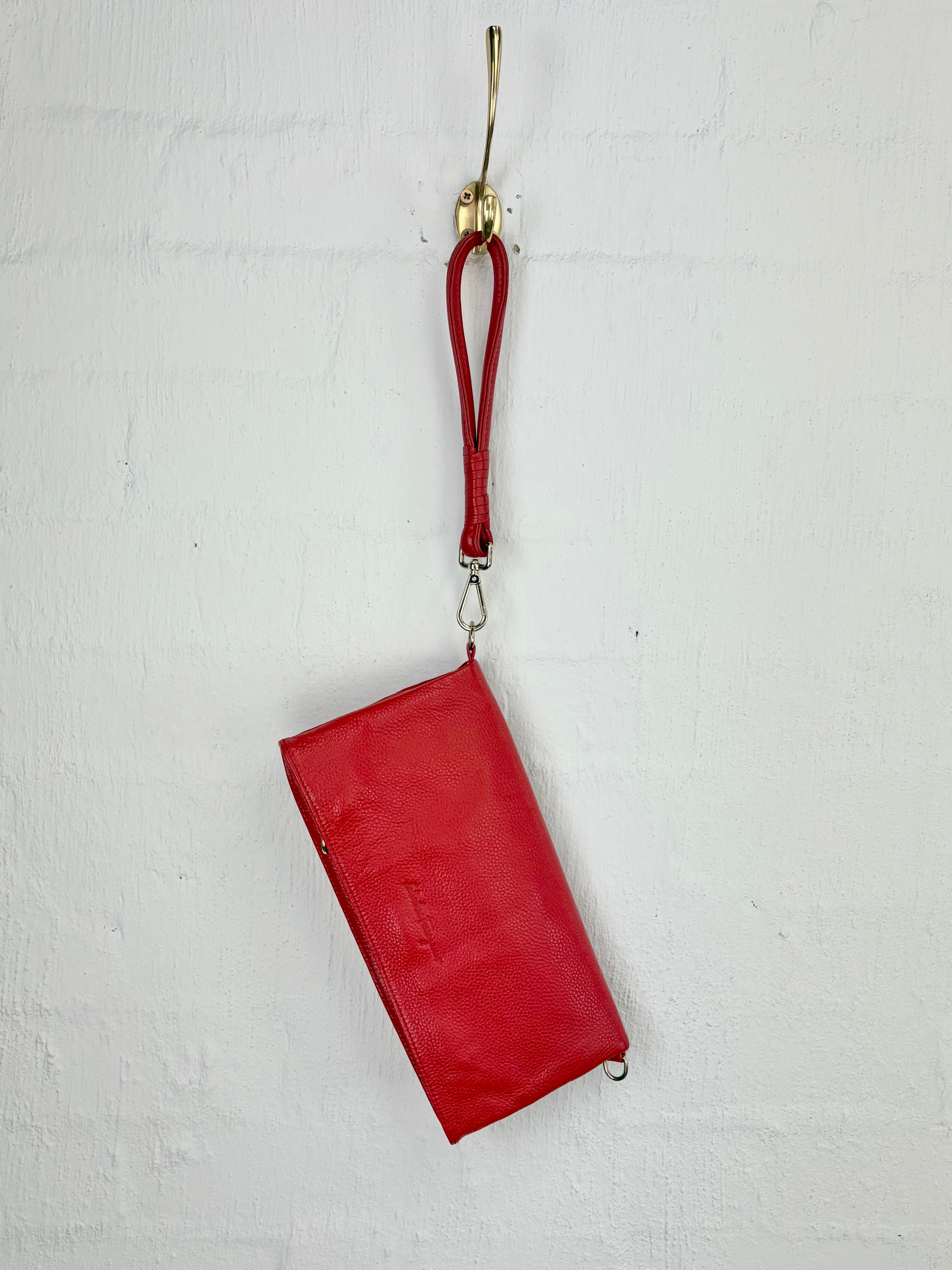 A red leather wristlet clutch bag with a matching red leather wrist strap, hanging from a gold metal hook against a white painted brick wall.