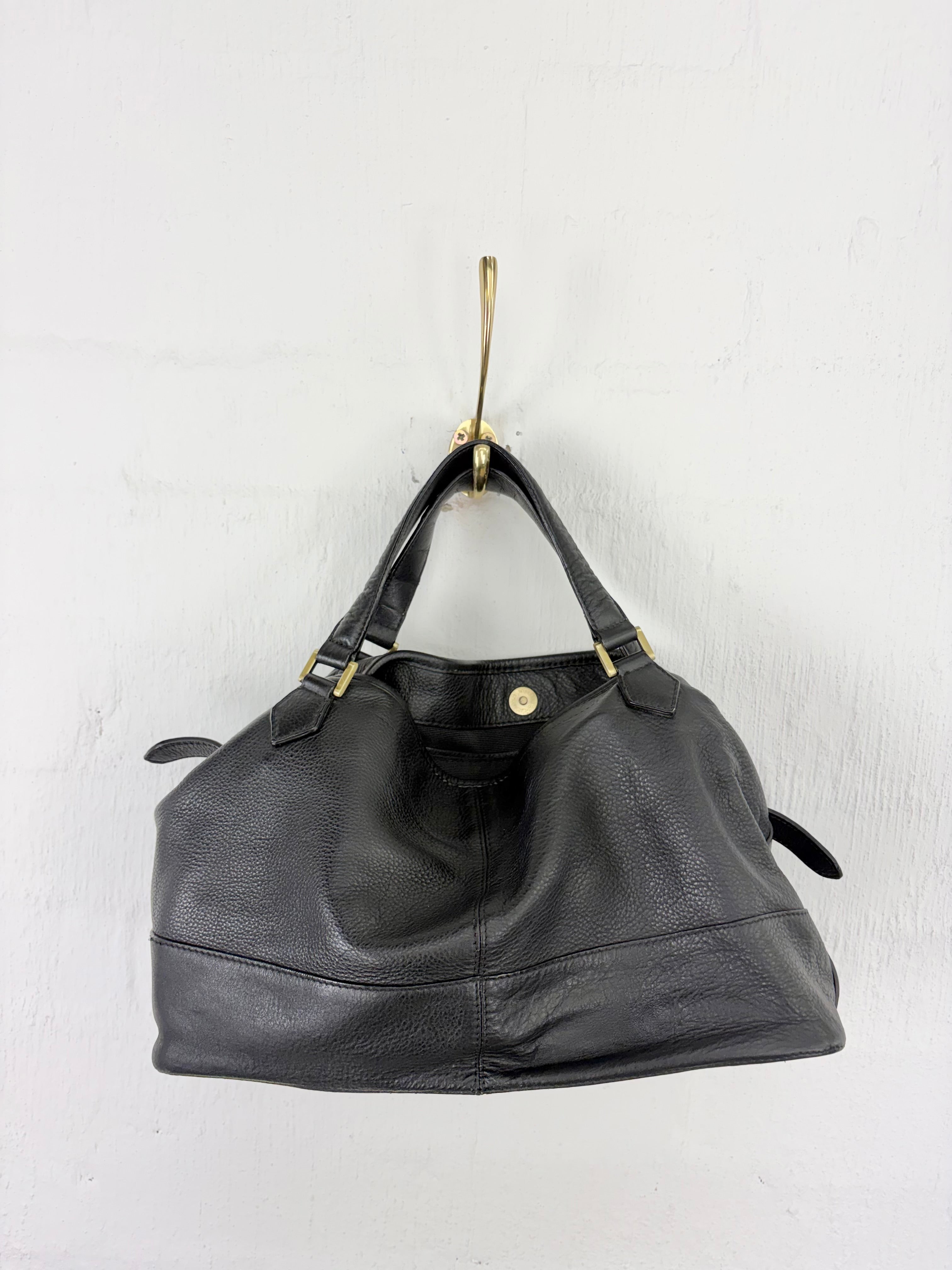A black pebbled leather handbag with gold-tone hardware, hanging from a gold hook against a white textured wall.