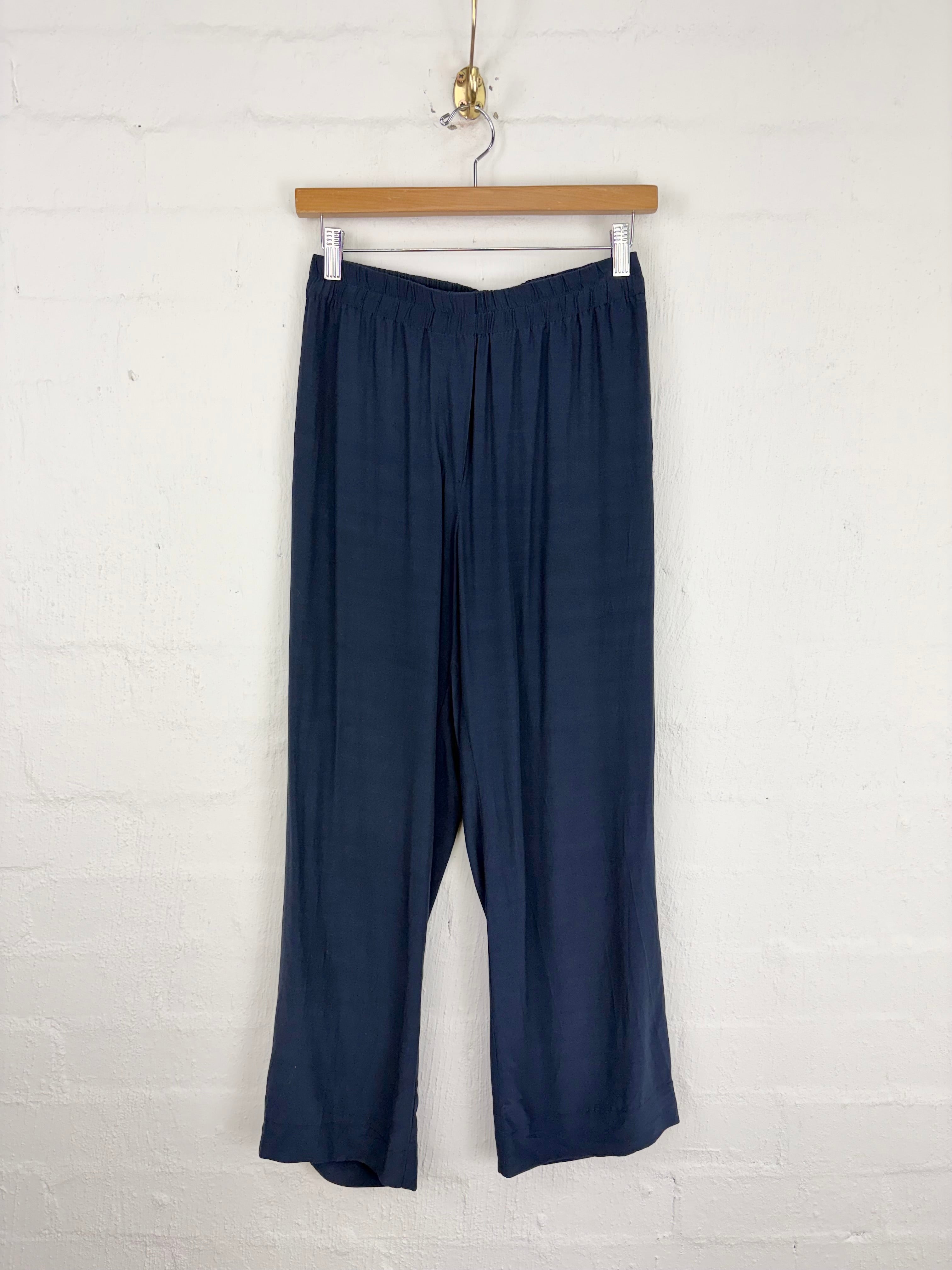 A pair of dark navy blue wide-leg pants hanging from a wooden hanger against a white painted brick wall.