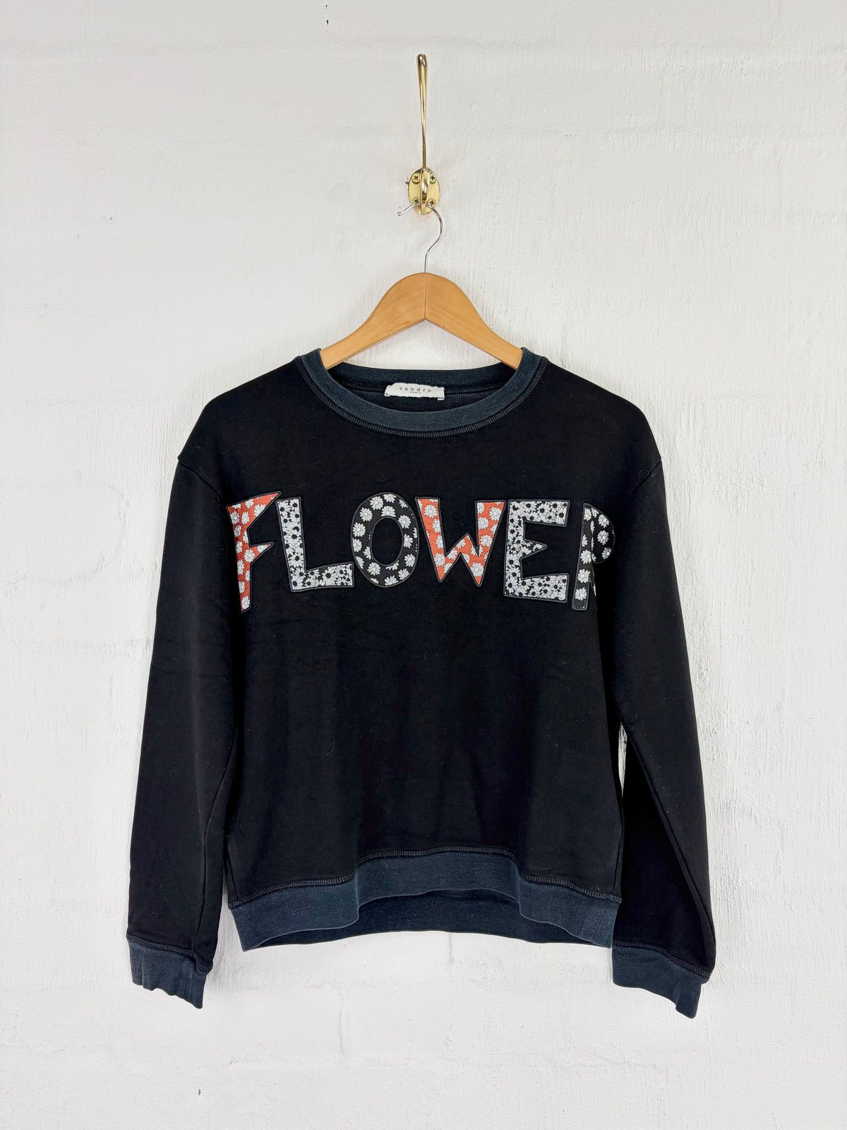 A black long-sleeve sweatshirt hanging on a wooden hanger against a white brick wall. The front features the word 'FLOWER' in large letters, with 'F' and 'E' filled with a red-and-white floral pattern, and 'W' and 'R' filled with white rhinestones.