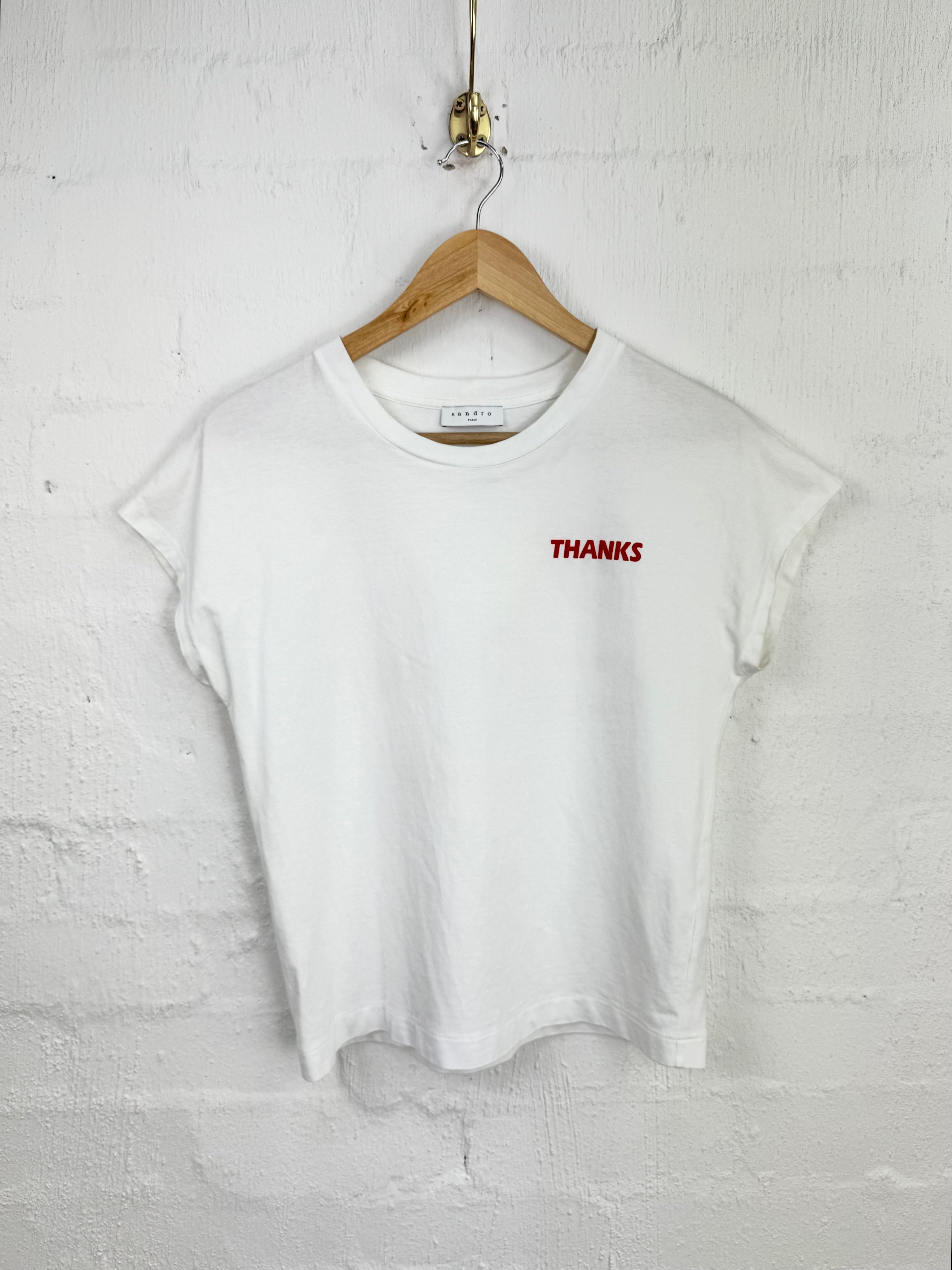 A white short-sleeve crop top t-shirt with 'THANKS' printed in red on the left chest, hanging on a wooden hanger against a white painted brick wall.