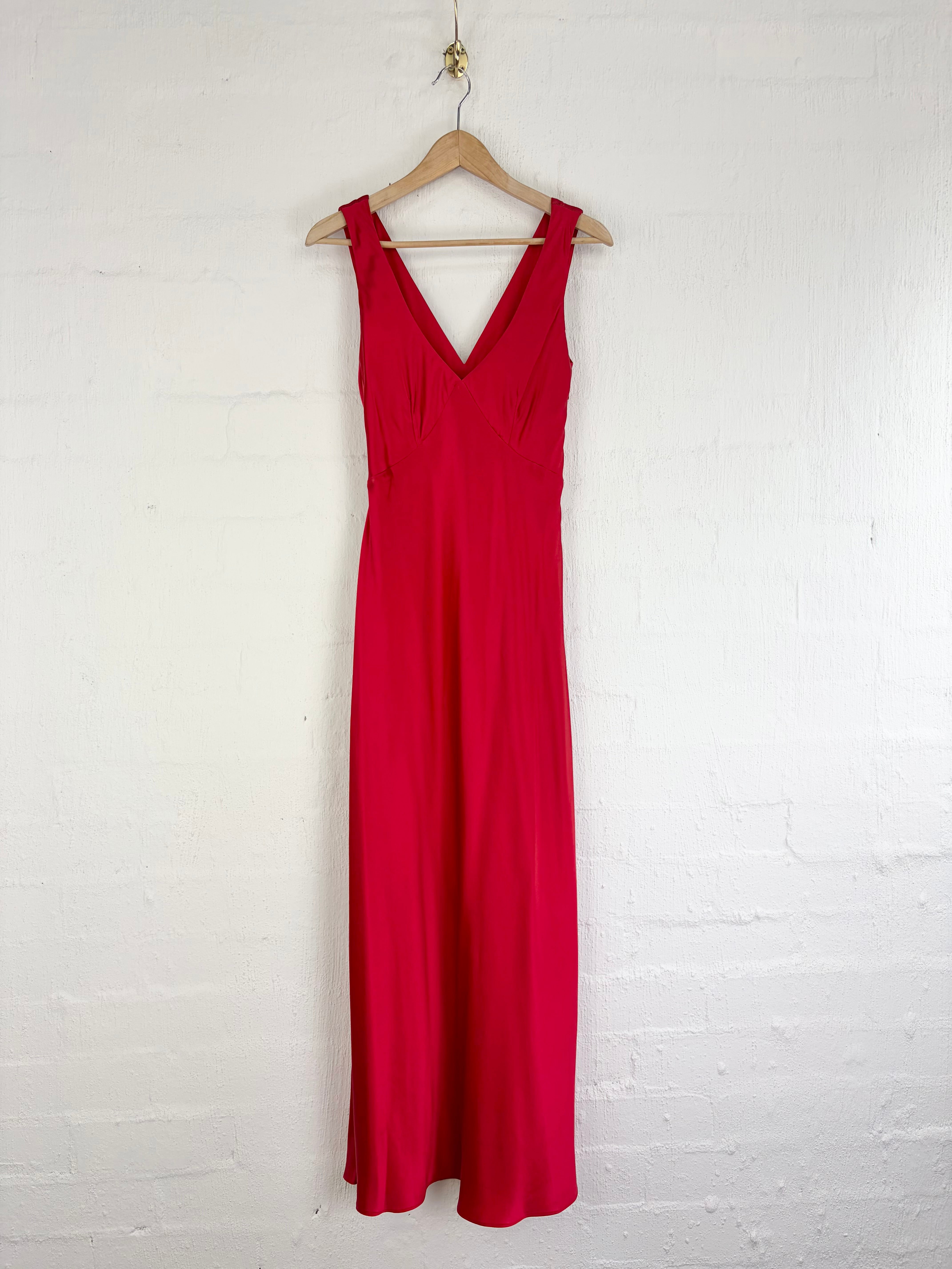 A bright red sleeveless maxi dress with a V-neckline, hanging on a wooden hanger against a white painted brick wall.