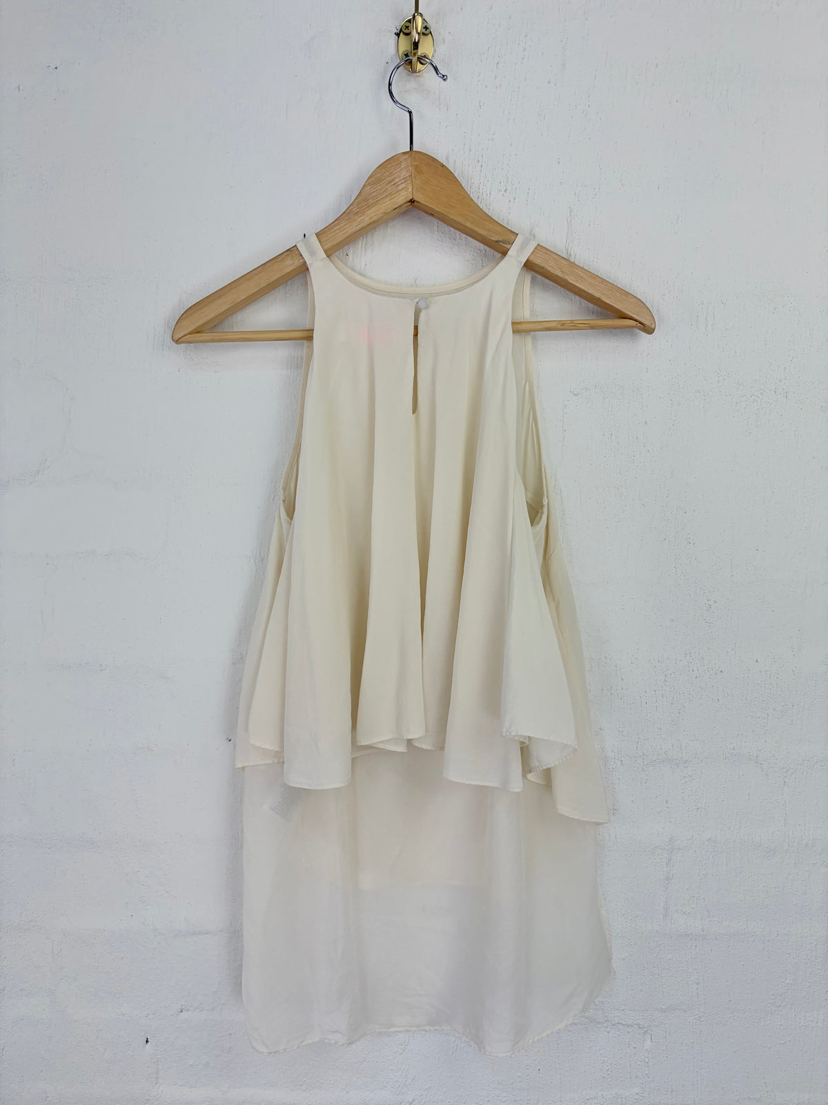 An off-white sleeveless blouse with a high-low layered skirt, hanging on a wooden hanger against a white painted brick wall.