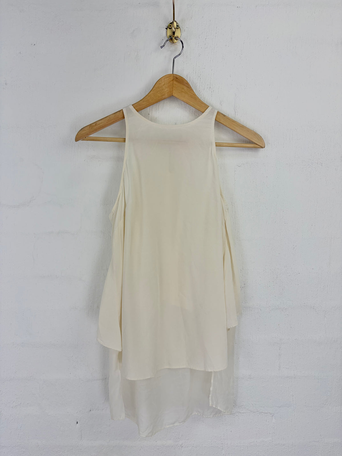 An off-white sleeveless high-low blouse hanging on a wooden hanger against a white painted brick wall.
