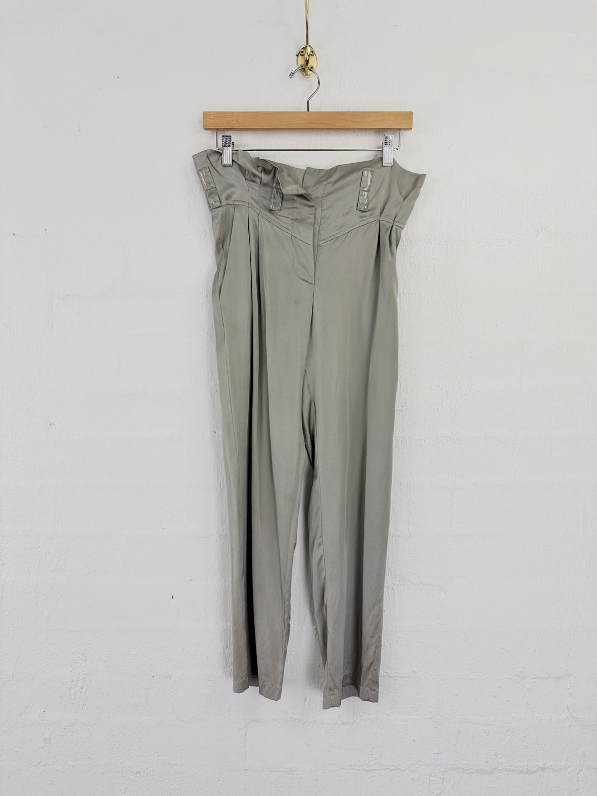 A pair of high-waisted, wide-leg trousers in a muted sage green or gray-green color, hanging from a wooden hanger against a white painted brick wall.