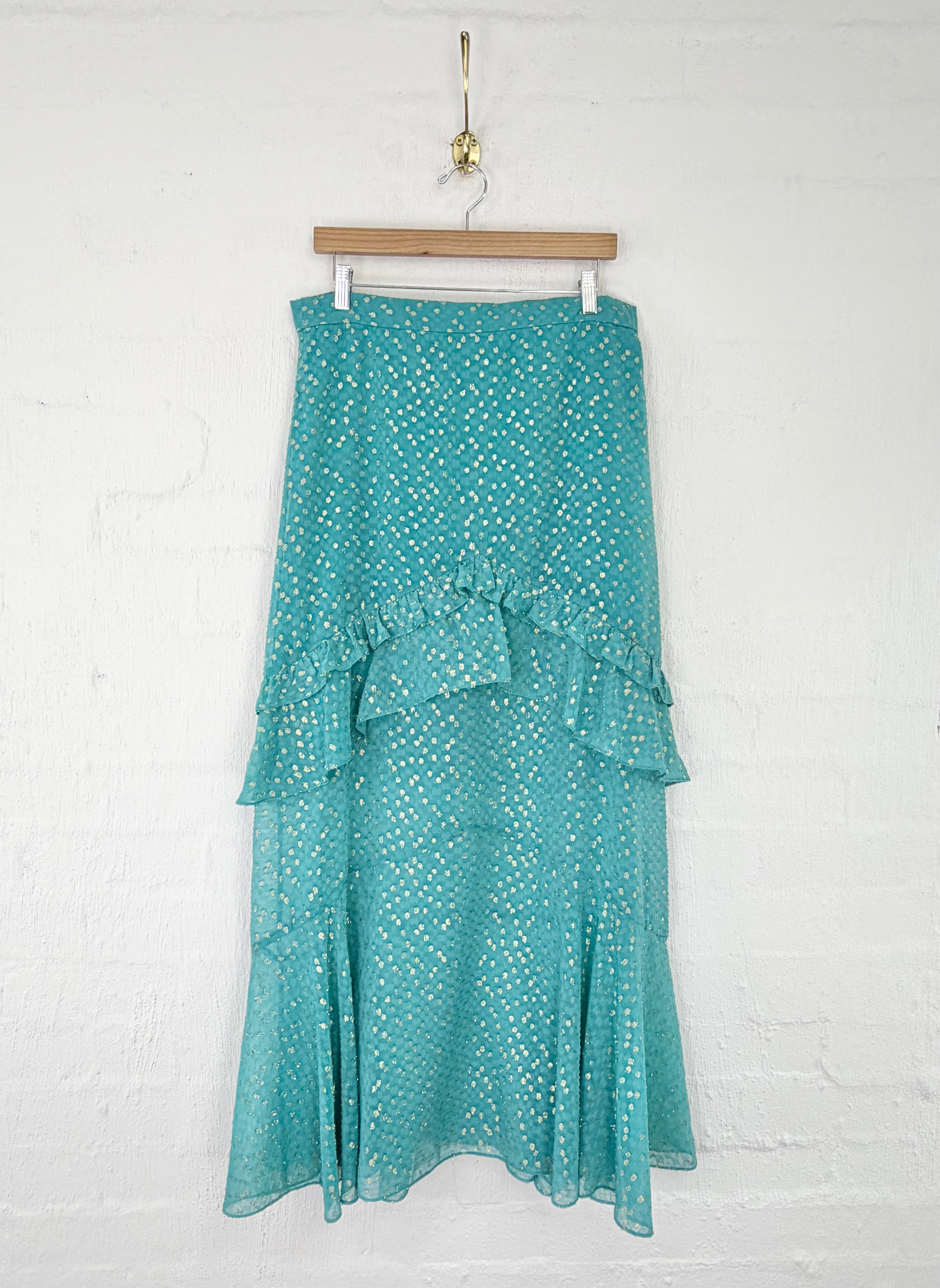 A teal blue tiered midi skirt with a white polka dot pattern, hanging from a wooden hanger against a white painted brick wall.