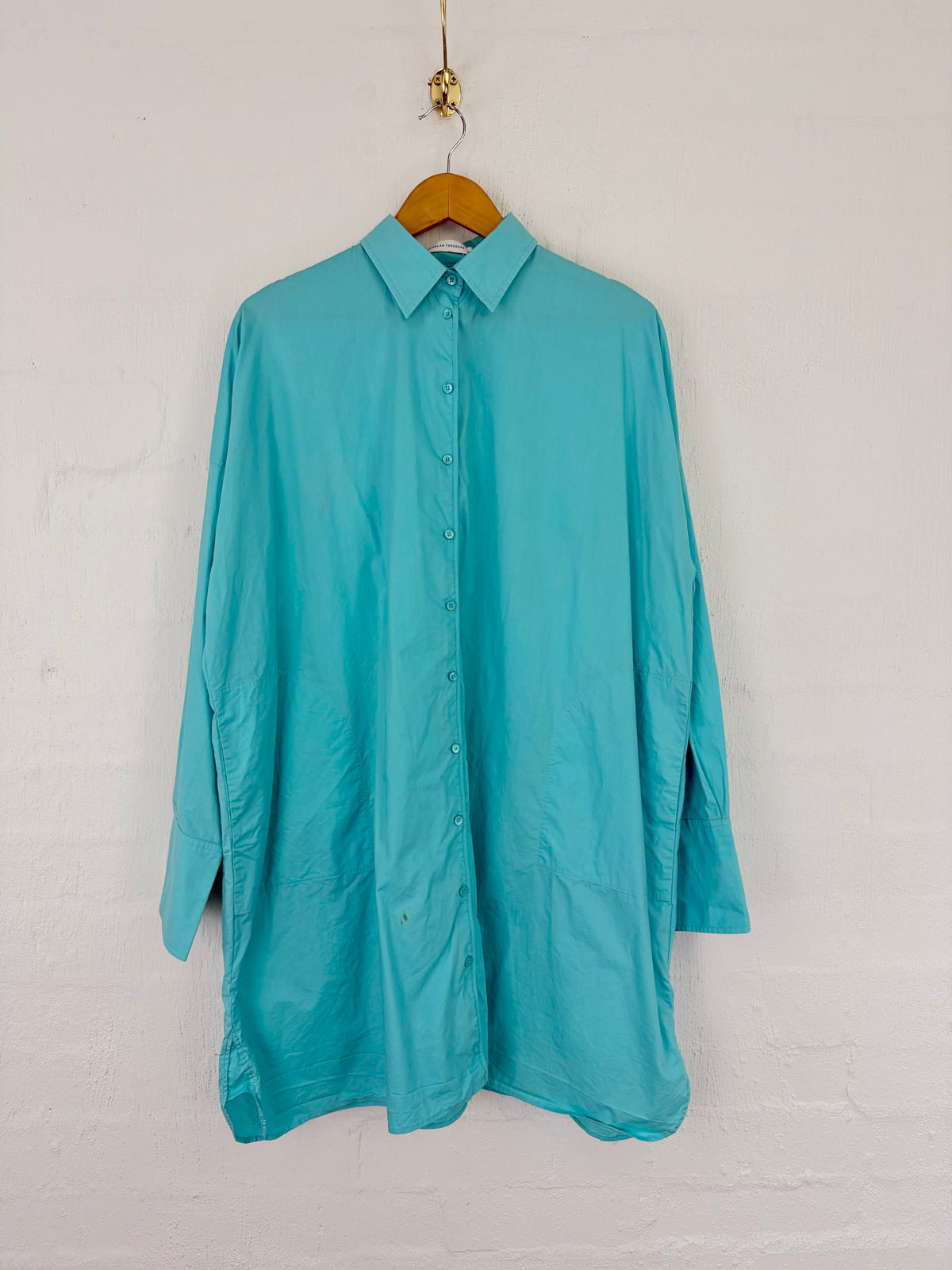 A turquoise long-sleeve button-up shirt hanging on a wooden hanger against a white painted brick wall.