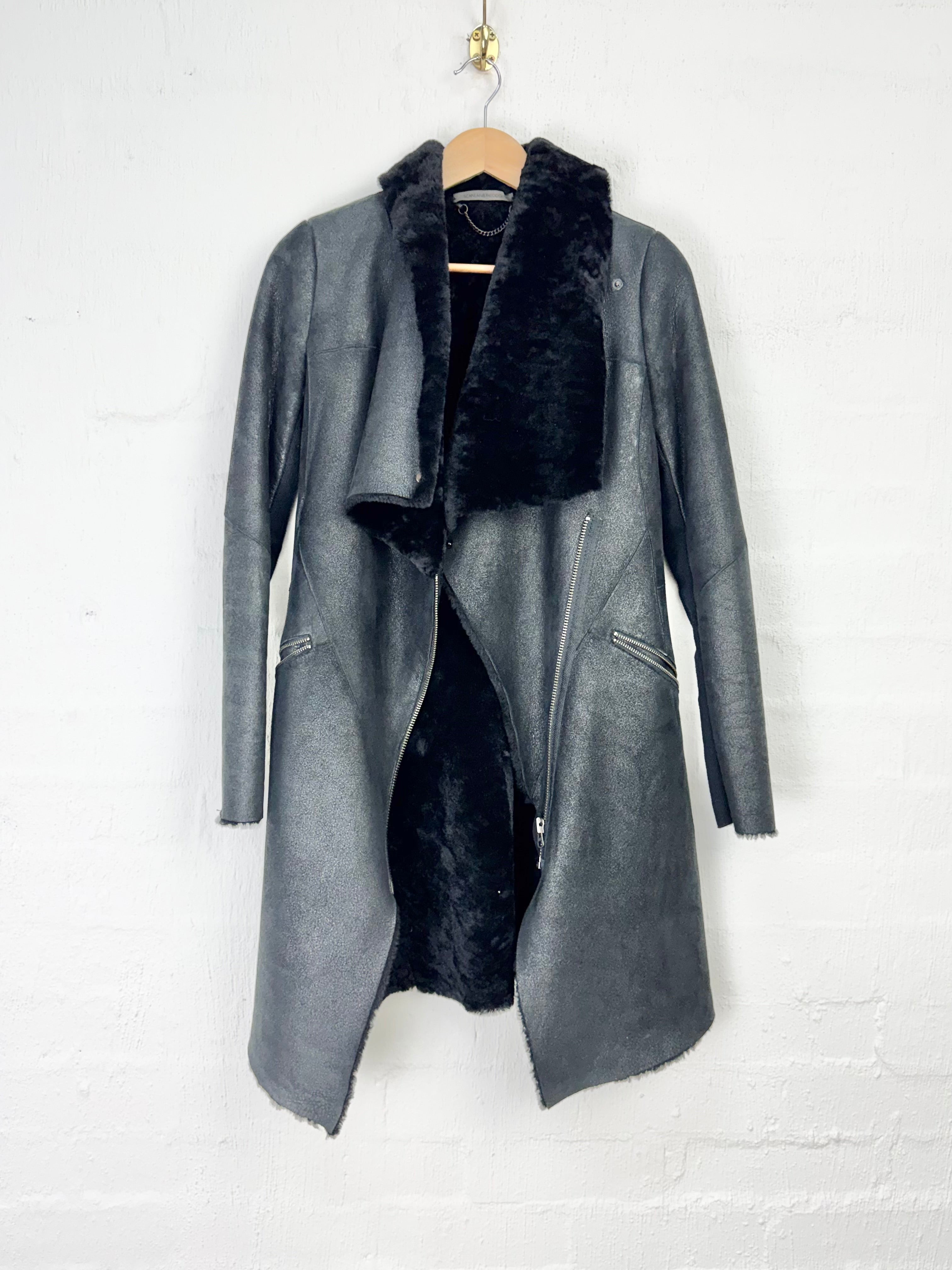 A dark gray faux-leather coat with a black faux-fur collar and lining, hanging on a wooden hanger against a white brick wall.