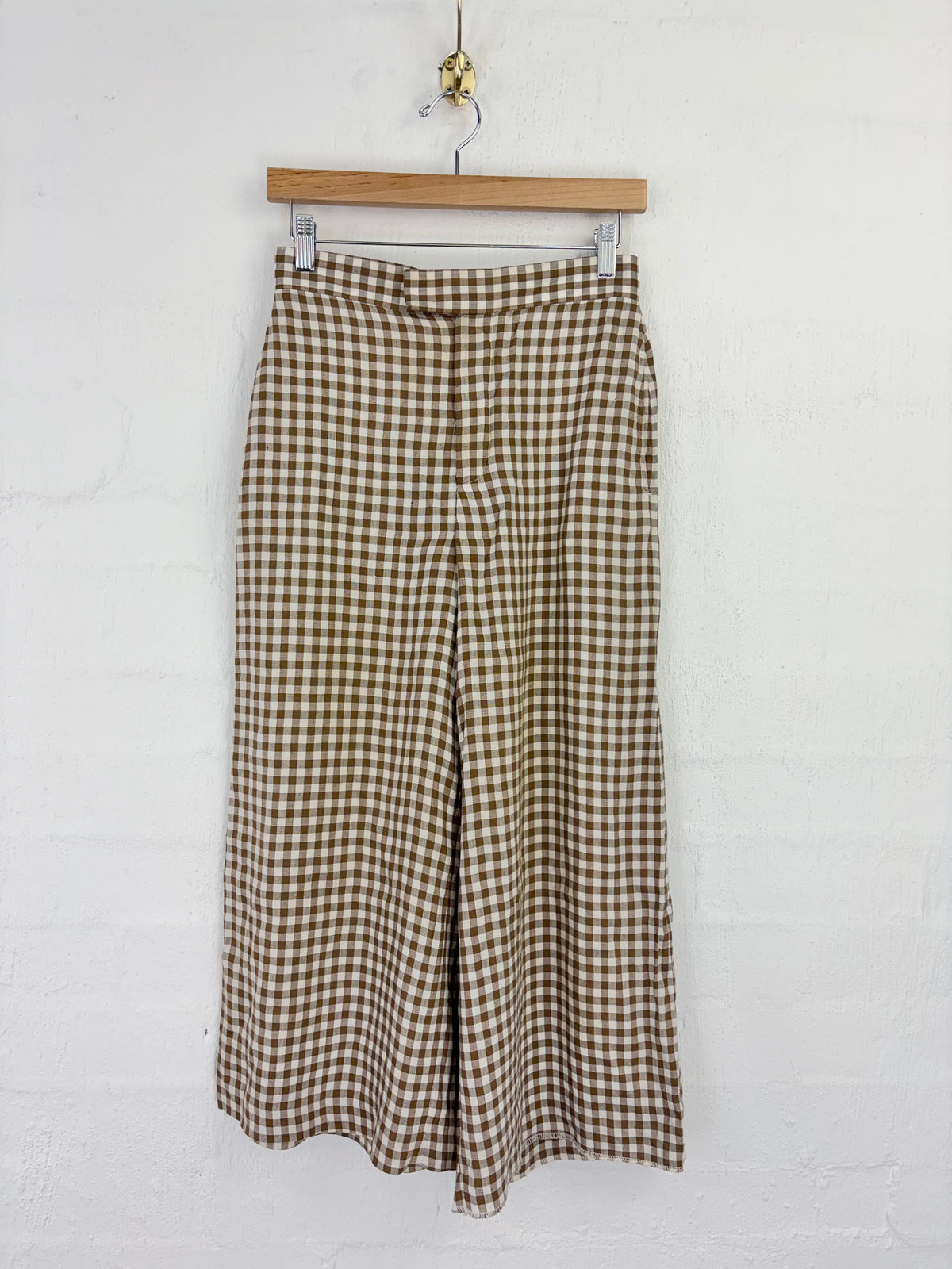 Brown and beige gingham cropped wide-leg pants hanging from a wooden hanger against a white brick wall.