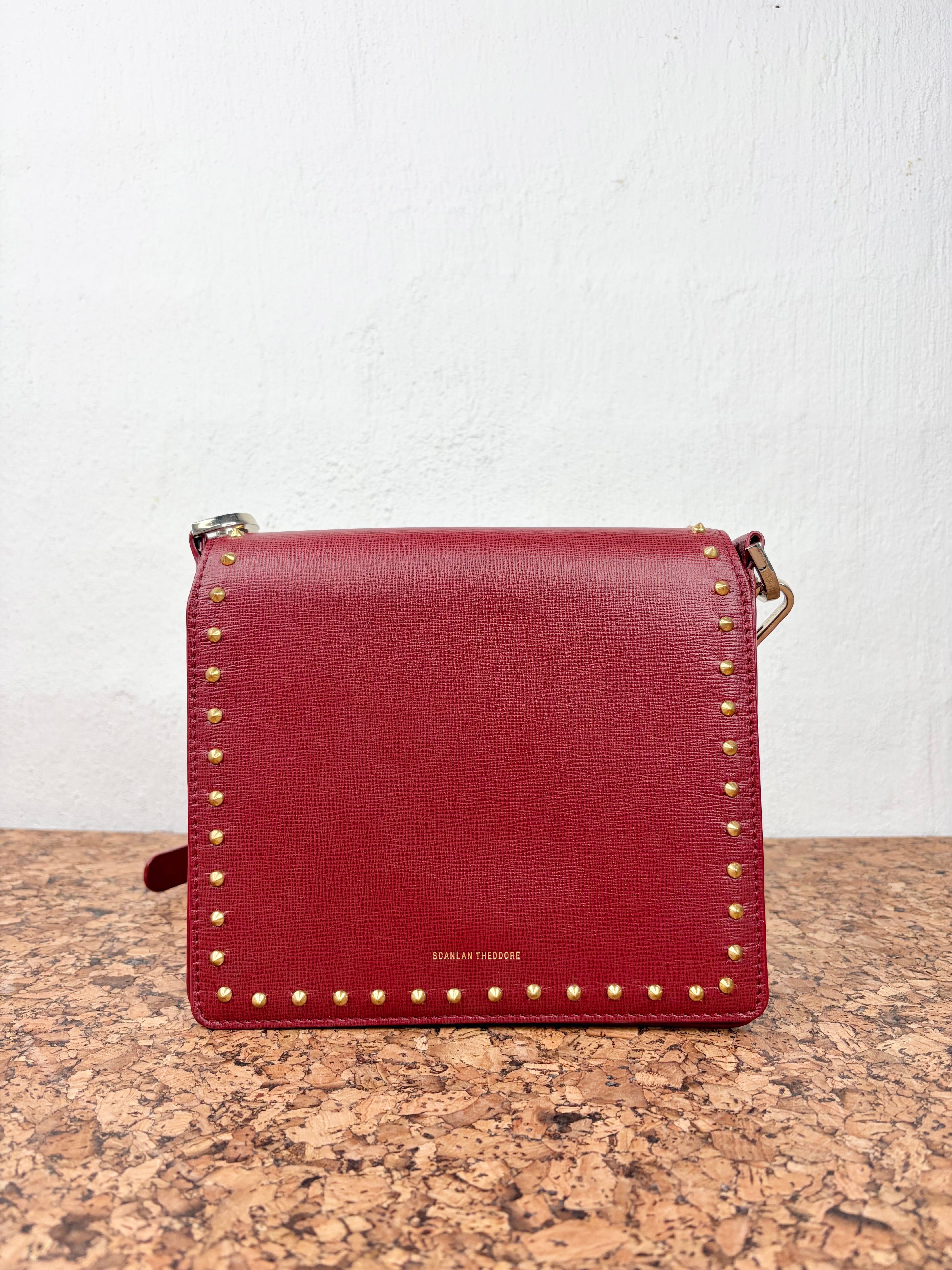 A red leather crossbody bag with gold-tone studs and hardware, displayed on a cork surface against a white wall.