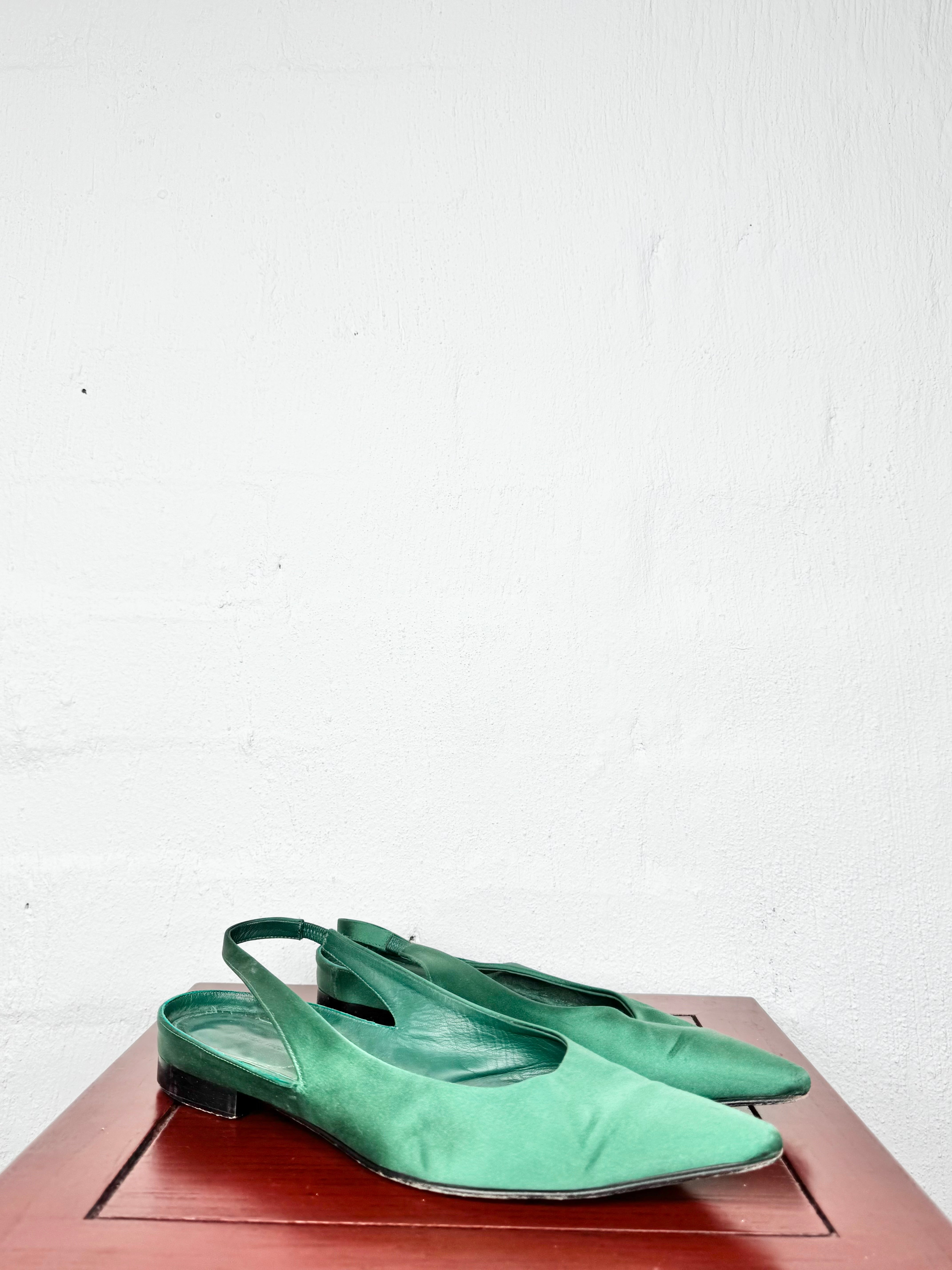 A pair of sage green satin slingback flats with pointed toes, displayed on a reddish-brown wooden surface against a white textured wall.