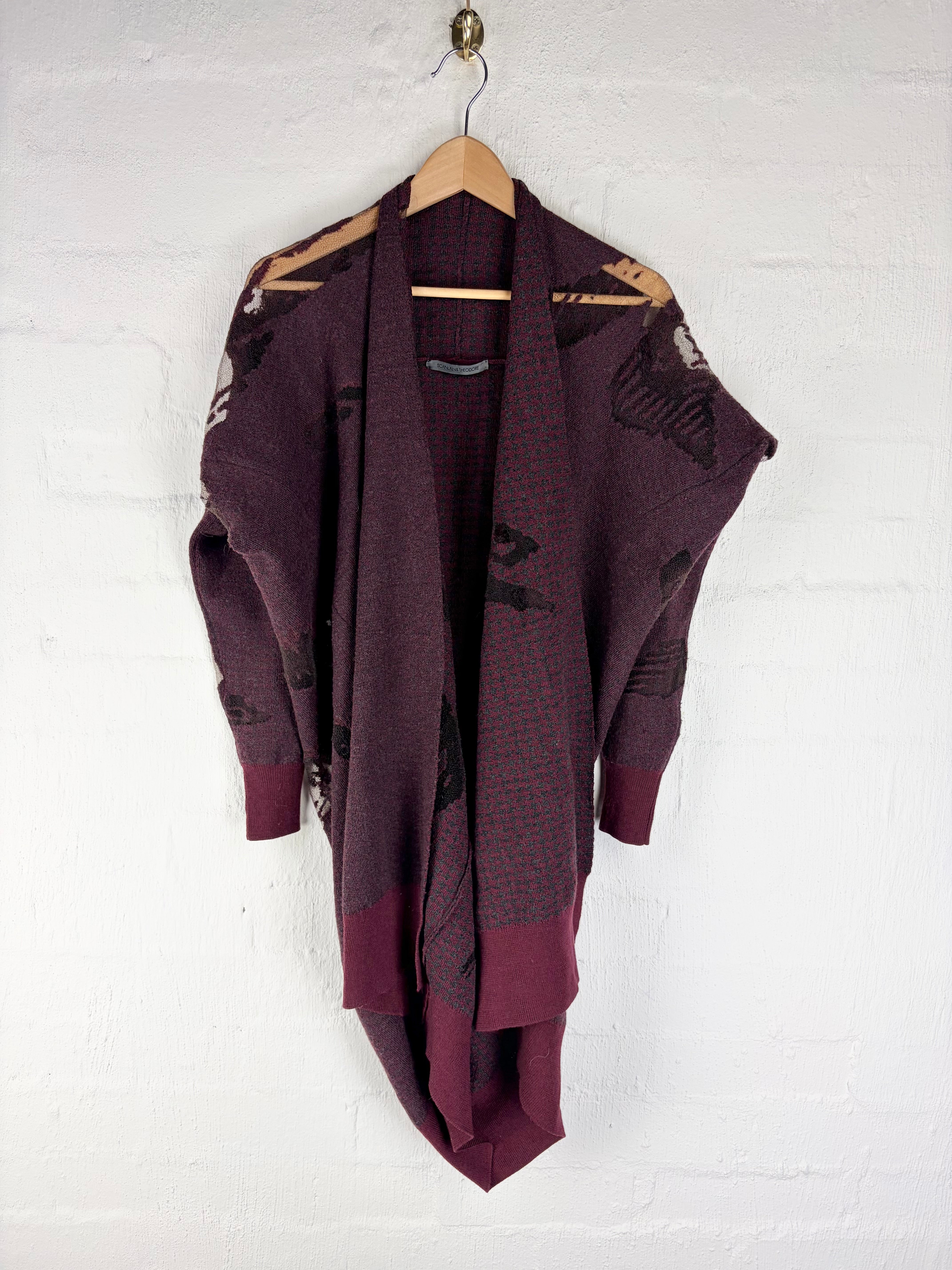 A dark plum purple knit cardigan with black abstract patterns, hanging on a wooden hanger against a white painted brick wall.