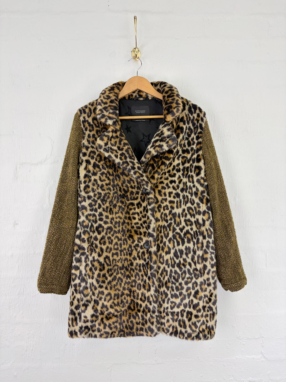 A women's coat hanging on a wooden hanger against a white painted brick wall. The coat has a plush leopard-print body in beige, brown, and black, paired with textured olive-brown sleeves.