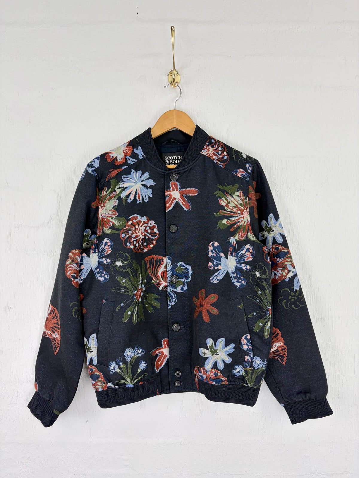 A black bomber jacket with a vibrant multicolor floral print, hanging on a wooden hanger against a white painted brick wall.