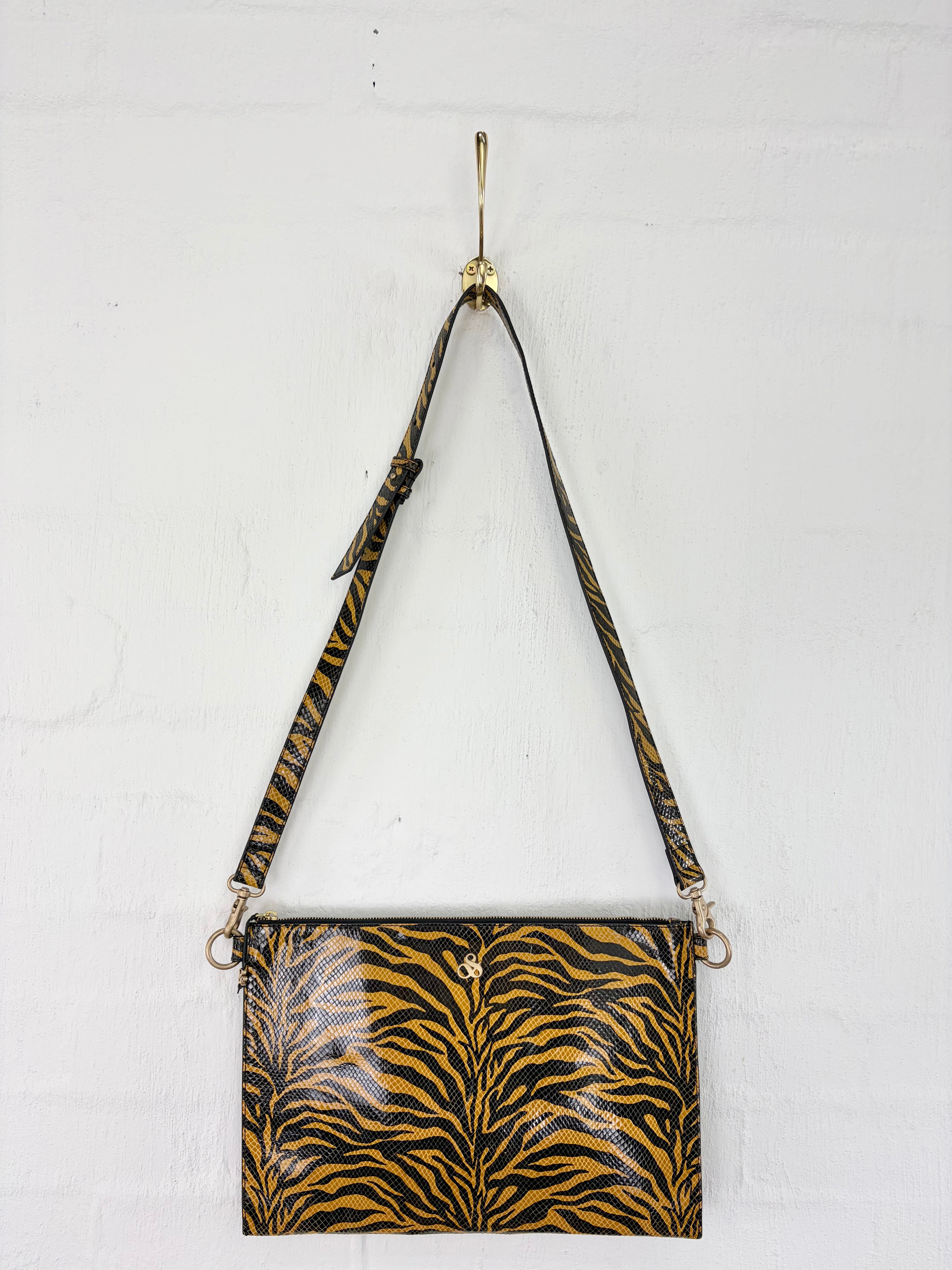 A rectangular shoulder bag with a glossy black and mustard yellow tiger-stripe pattern, hanging from a gold hook on a white brick wall.