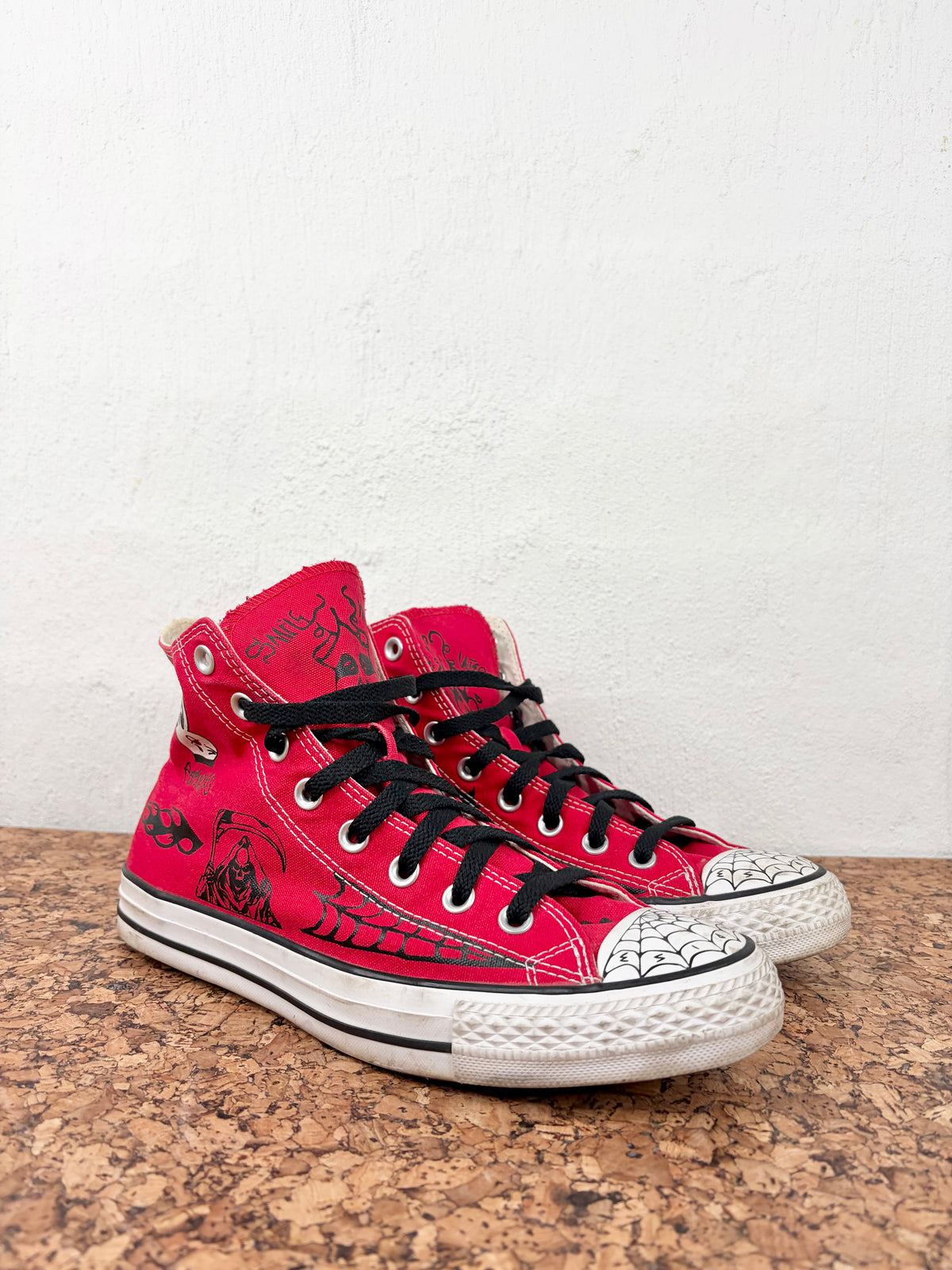 A pair of red high-top sneakers with black graphic prints, black laces, and white rubber toe caps, displayed on a cork surface against a white wall.