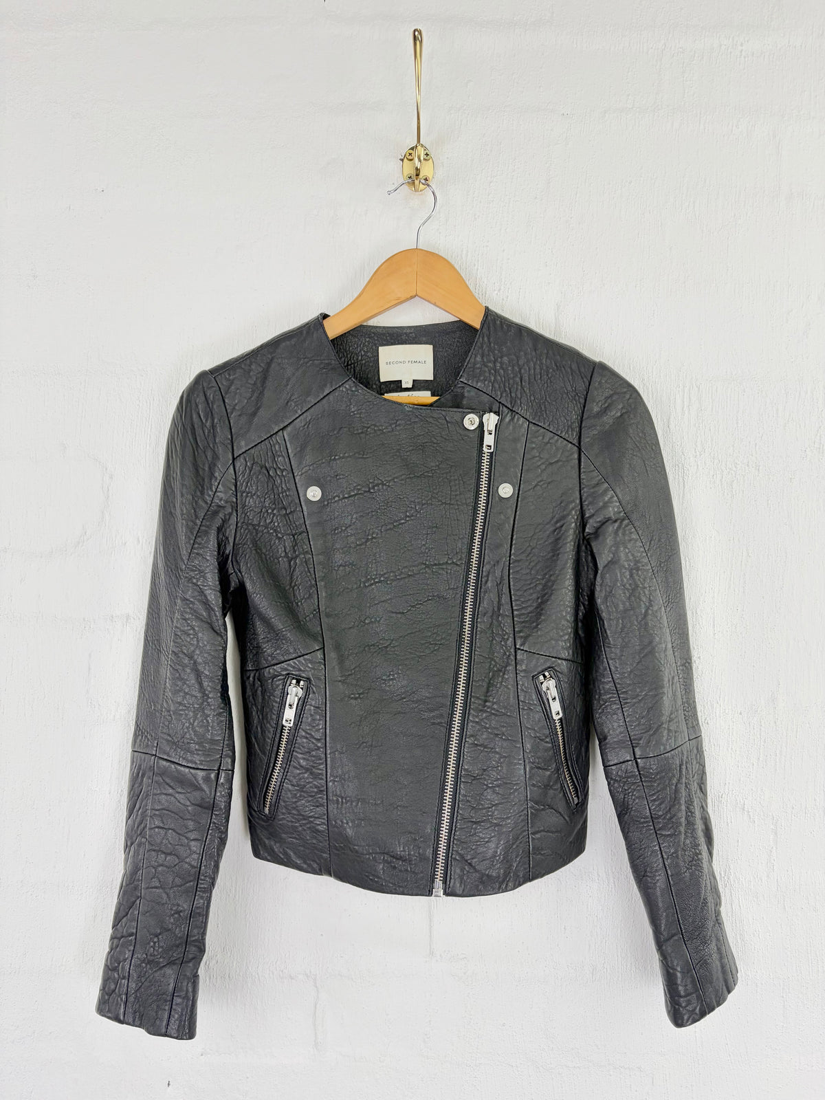 A dark gray faux-leather moto jacket with a textured crinkle finish, hanging on a wooden hanger against a white brick wall.