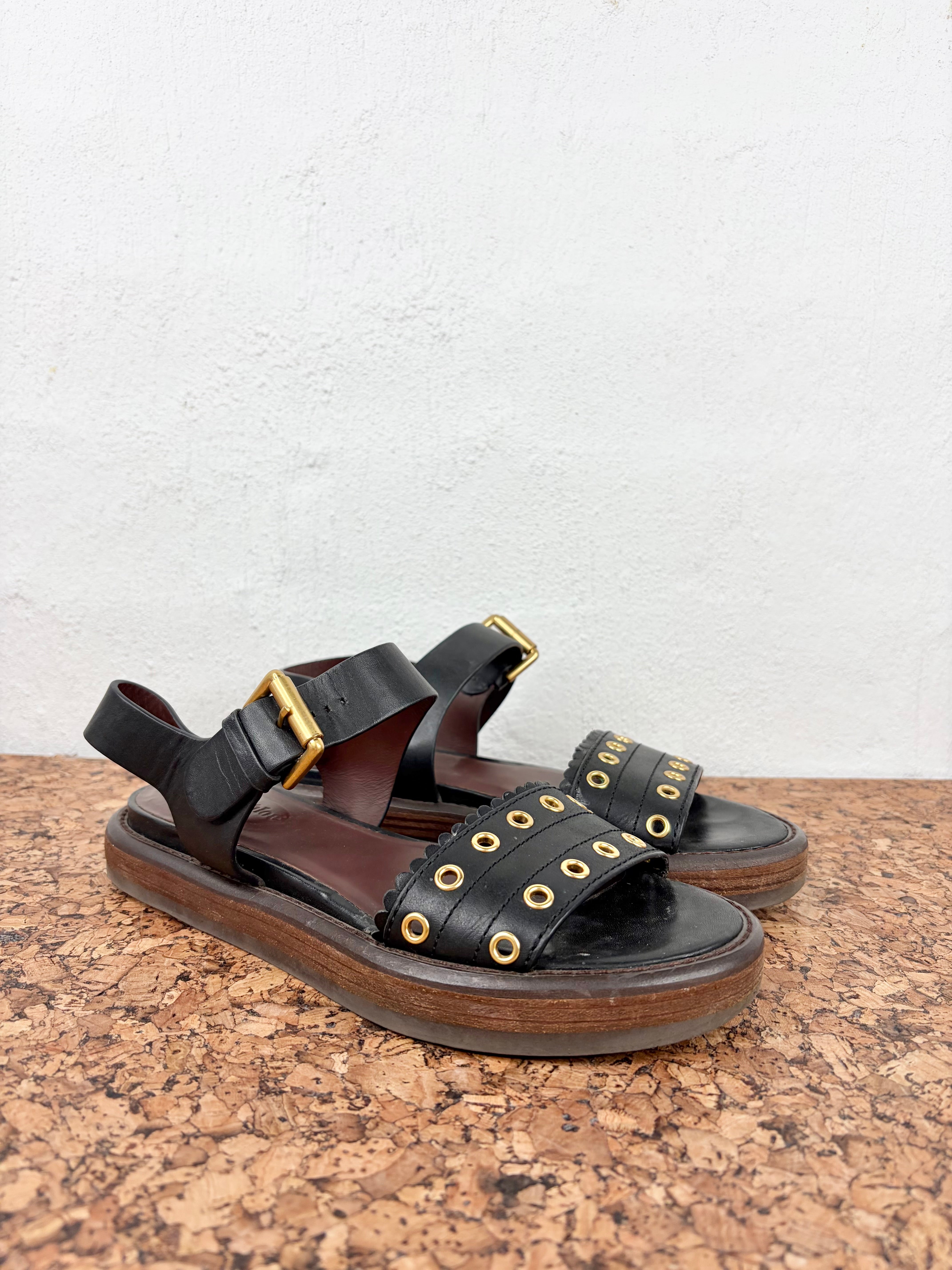 A pair of black leather platform sandals with gold-tone metal grommets and buckles, resting on a cork surface against a white wall.