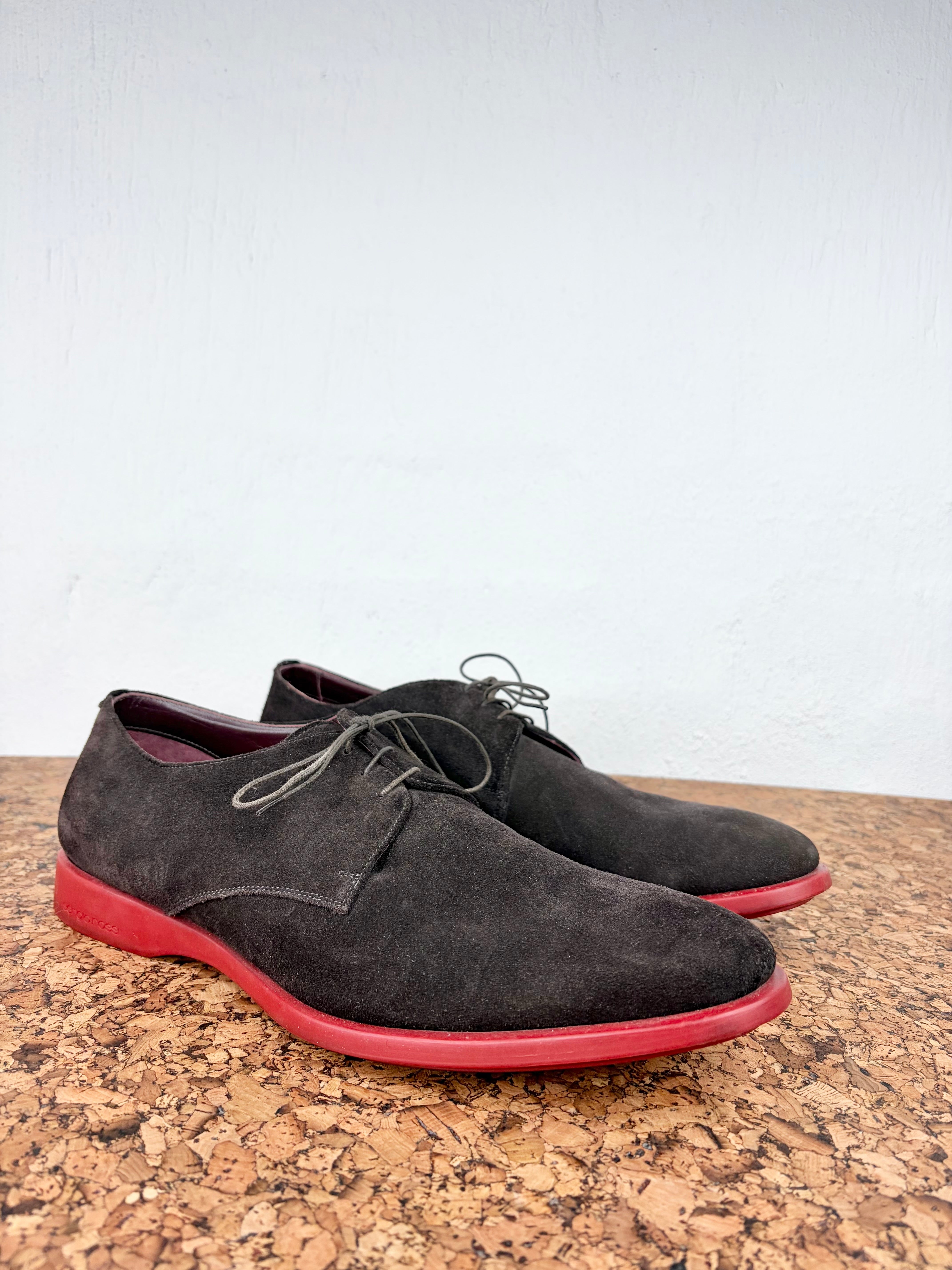 A pair of dark brown suede derby shoes with bright red soles, displayed on a cork surface against a white wall.