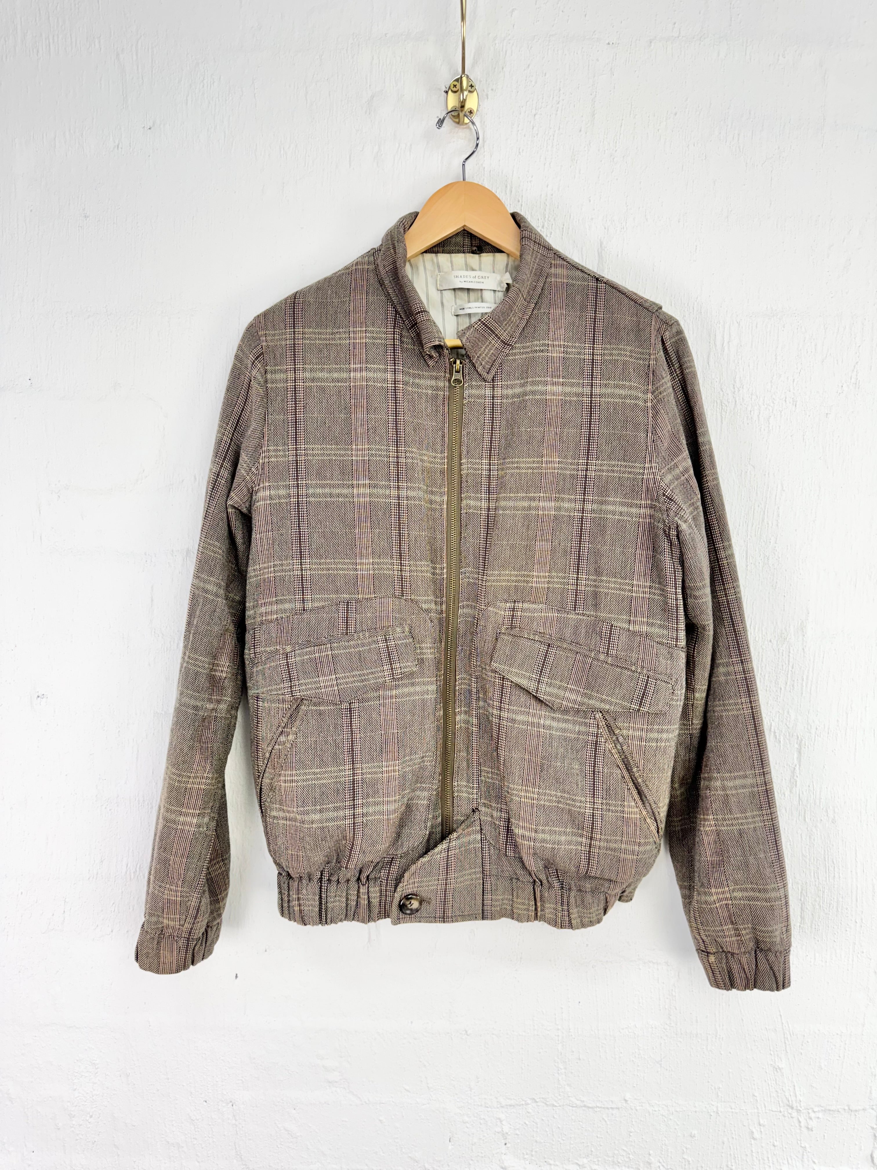 A vintage plaid zip-up jacket in brown and beige tones, hanging on a wooden hanger against a white painted brick wall.