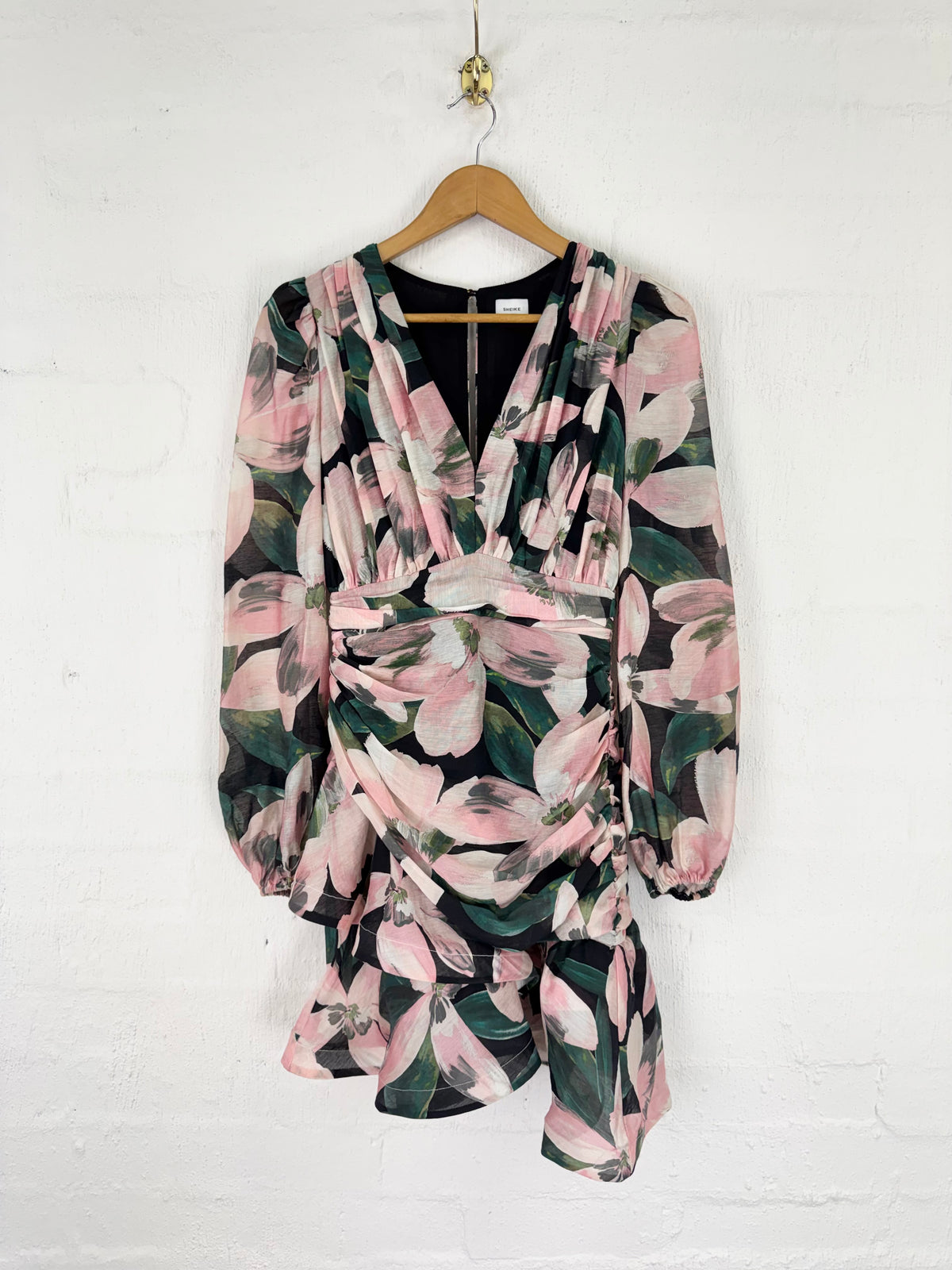 A long-sleeve wrap dress with a pink and green floral print, hanging on a wooden hanger against a white painted brick wall.
