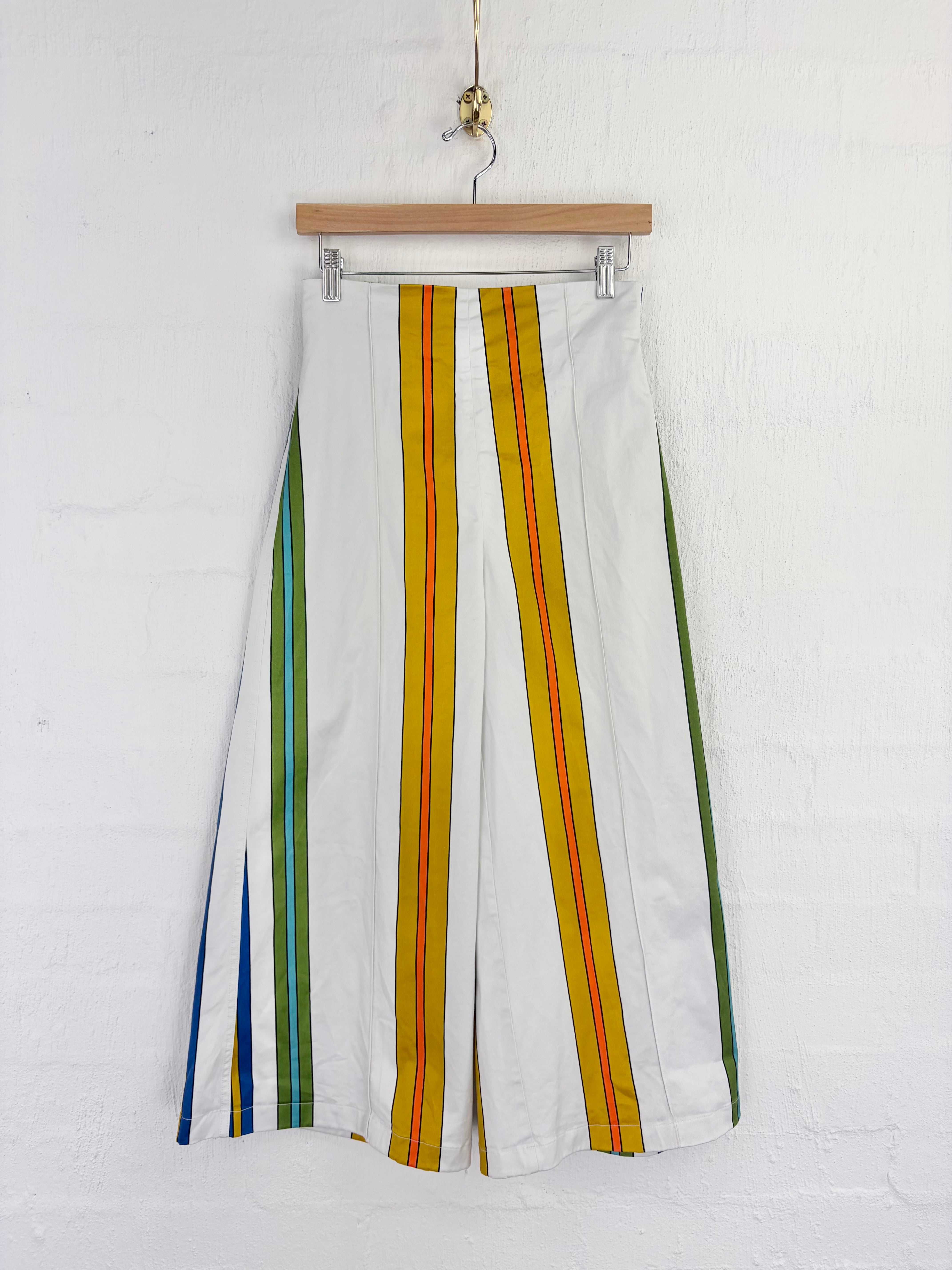 White wide-leg cropped pants with vertical multicolor stripes (yellow, orange, green, blue) hanging from a wooden hanger against a white brick wall.
