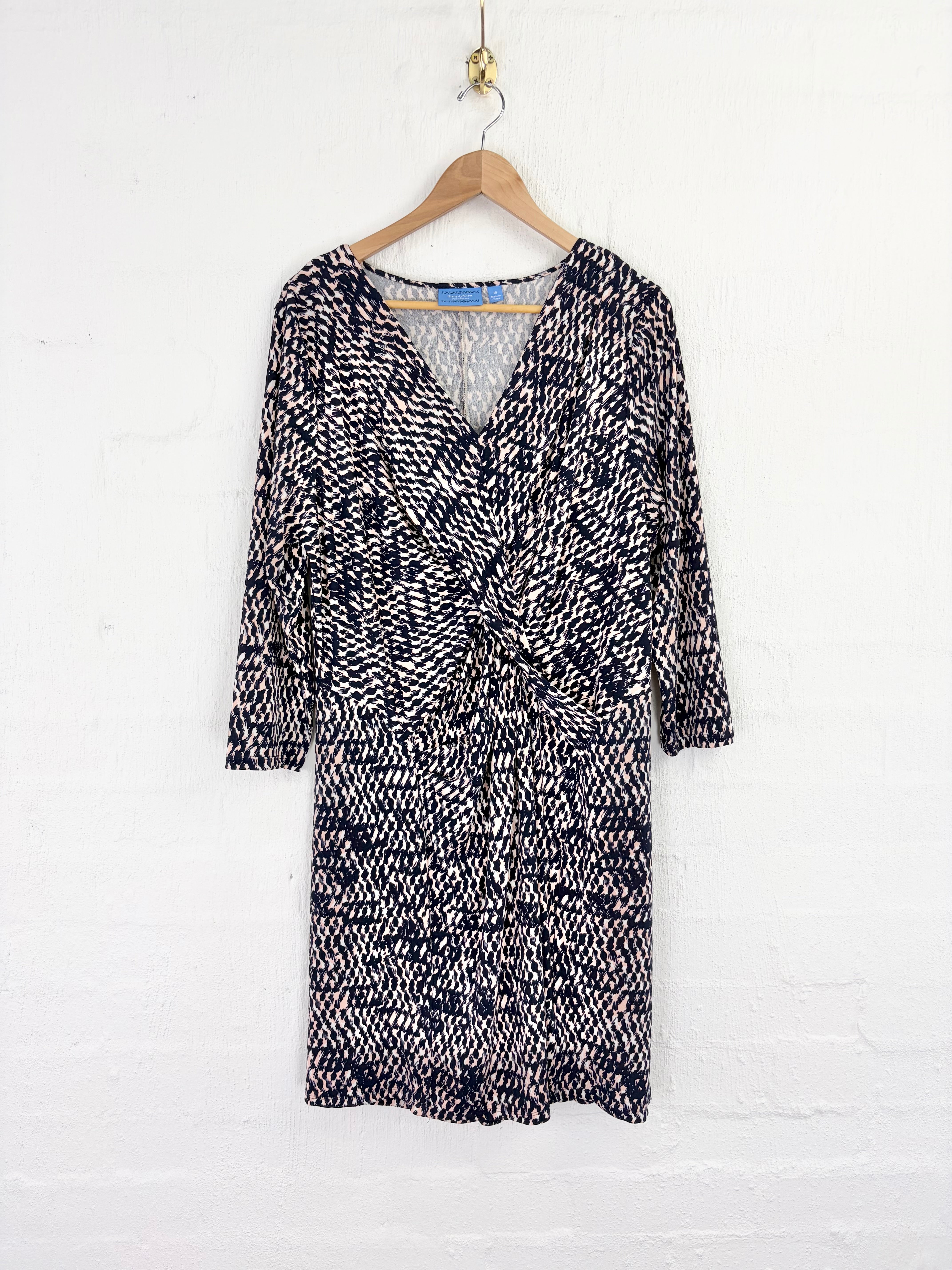 A black and white patterned wrap dress with three-quarter sleeves, hanging on a wooden hanger against a white brick wall.