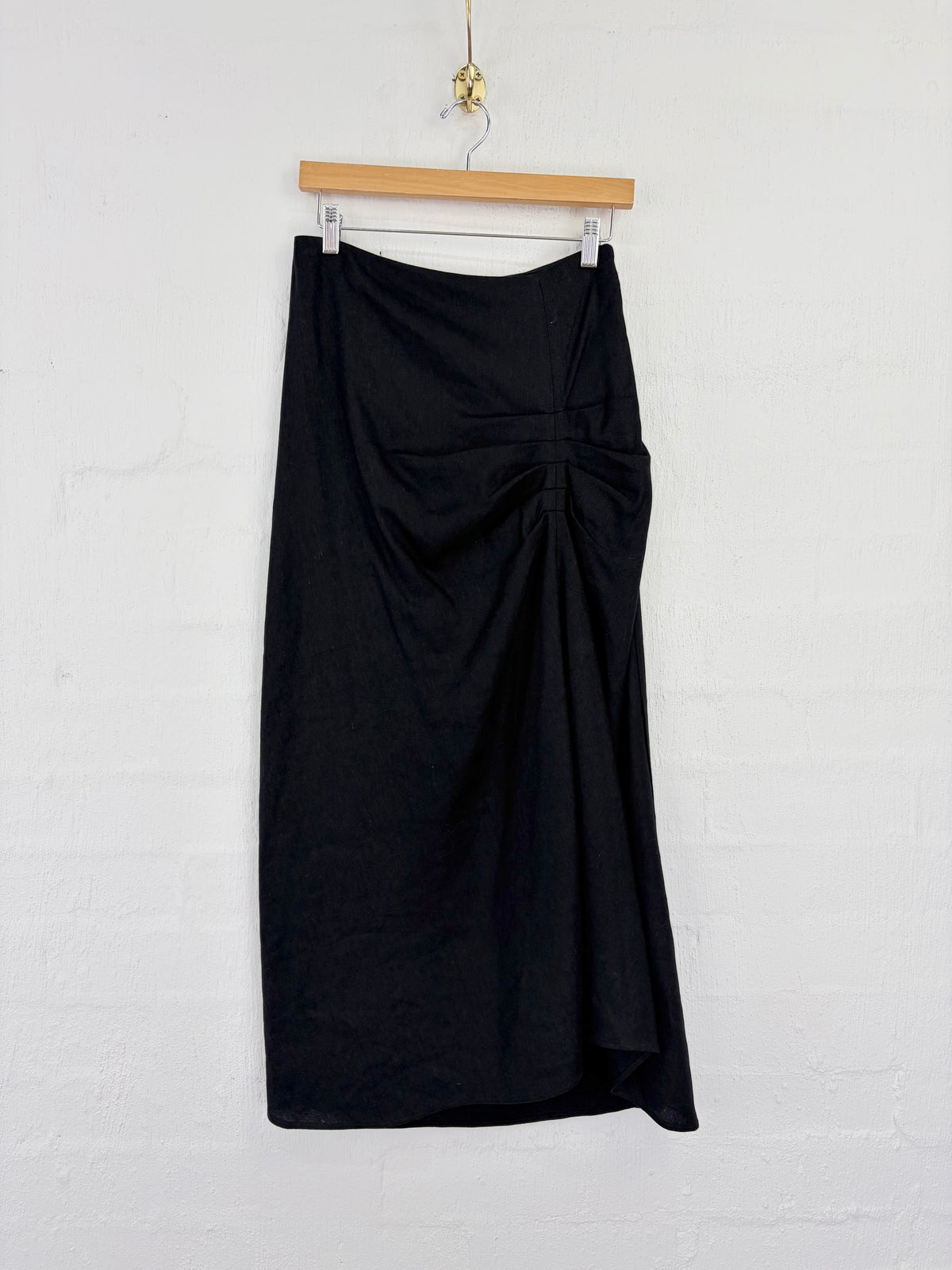 A black midi skirt with a gathered ruched front detail, hanging from a wooden hanger against a white painted brick wall.
