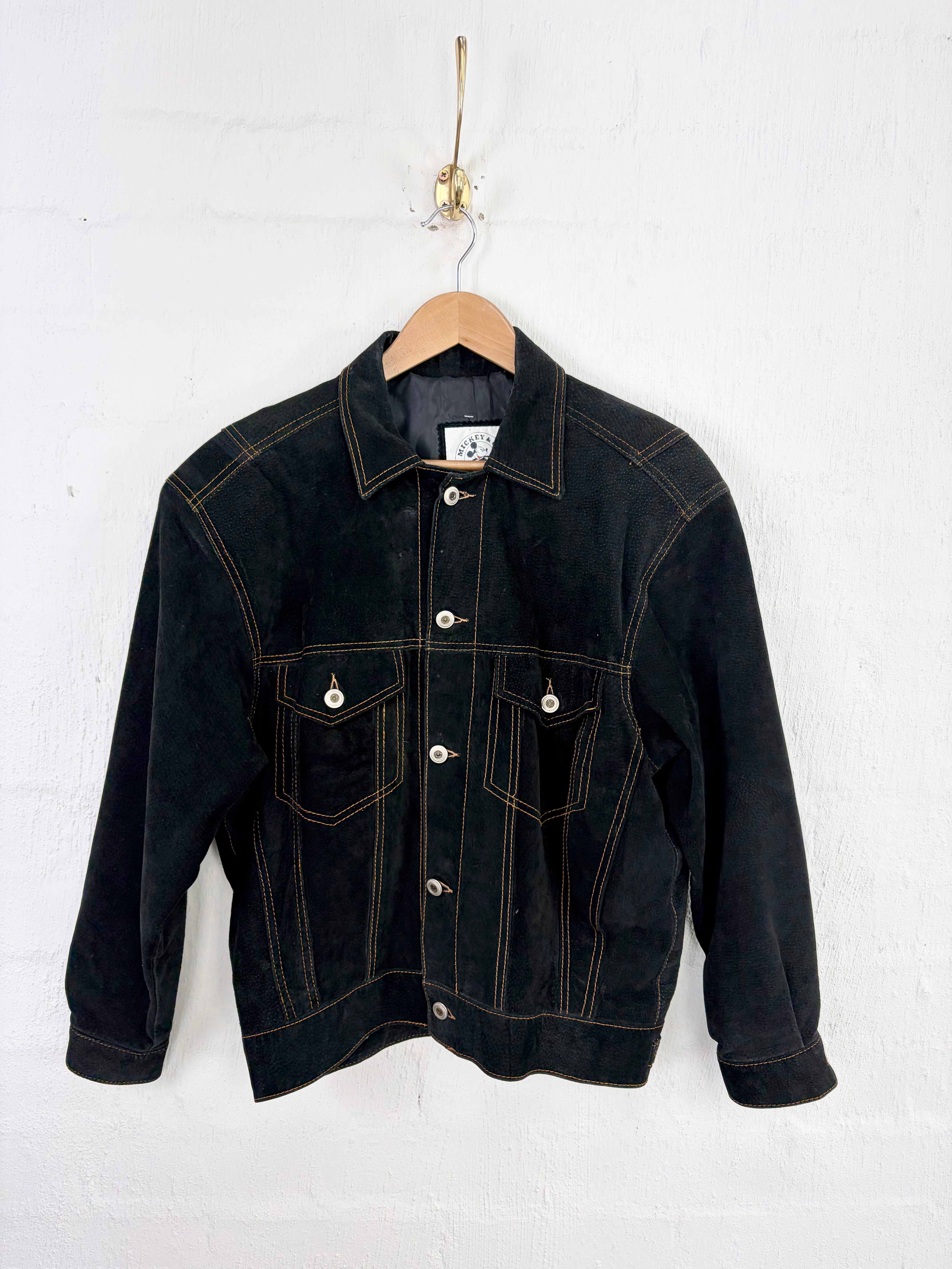 A black denim jacket with contrasting tan stitching, hanging on a wooden hanger against a white painted brick wall.