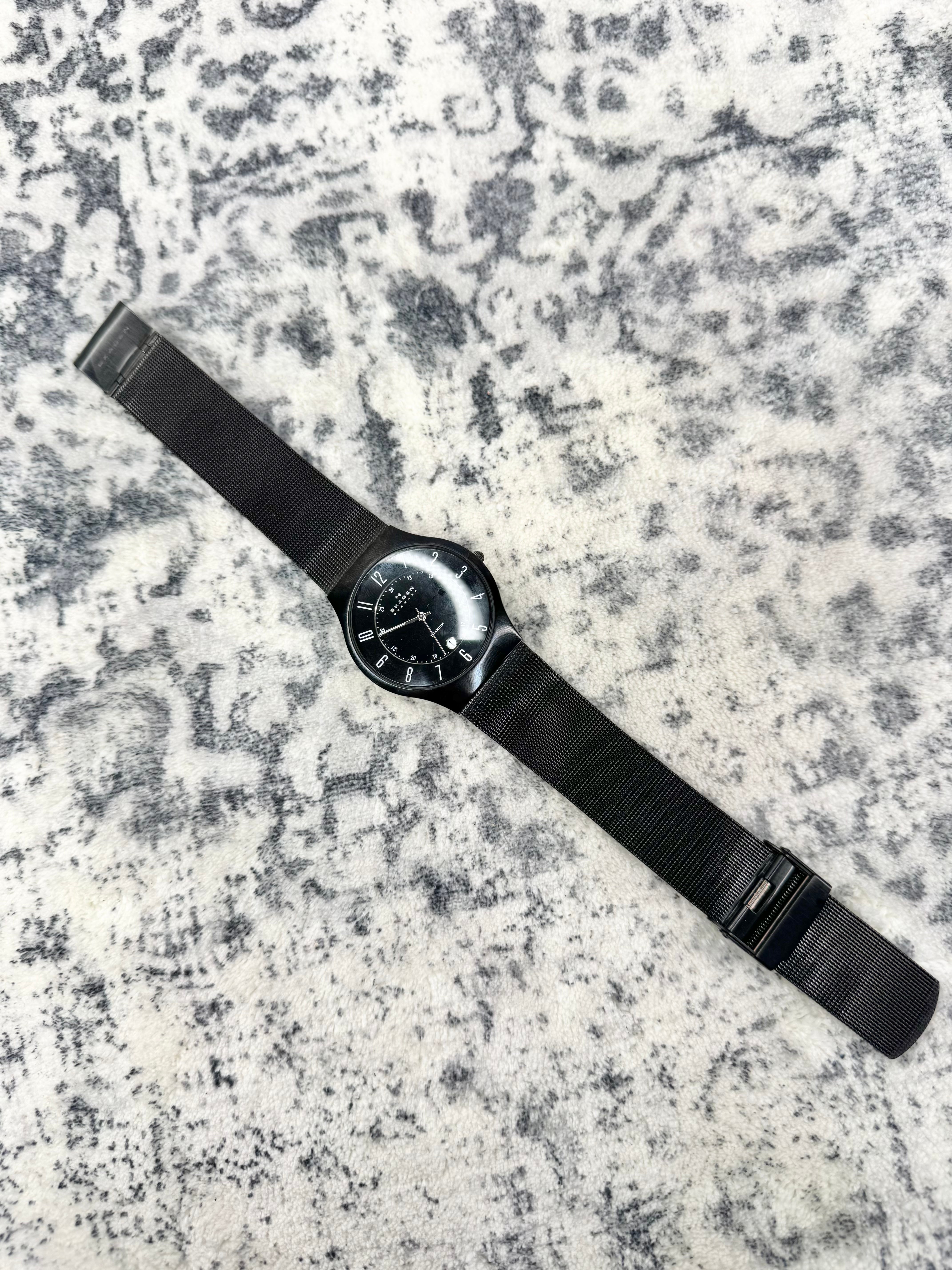 A black analog watch with a black dial and black mesh strap, laid diagonally on a textured gray and white stone surface.