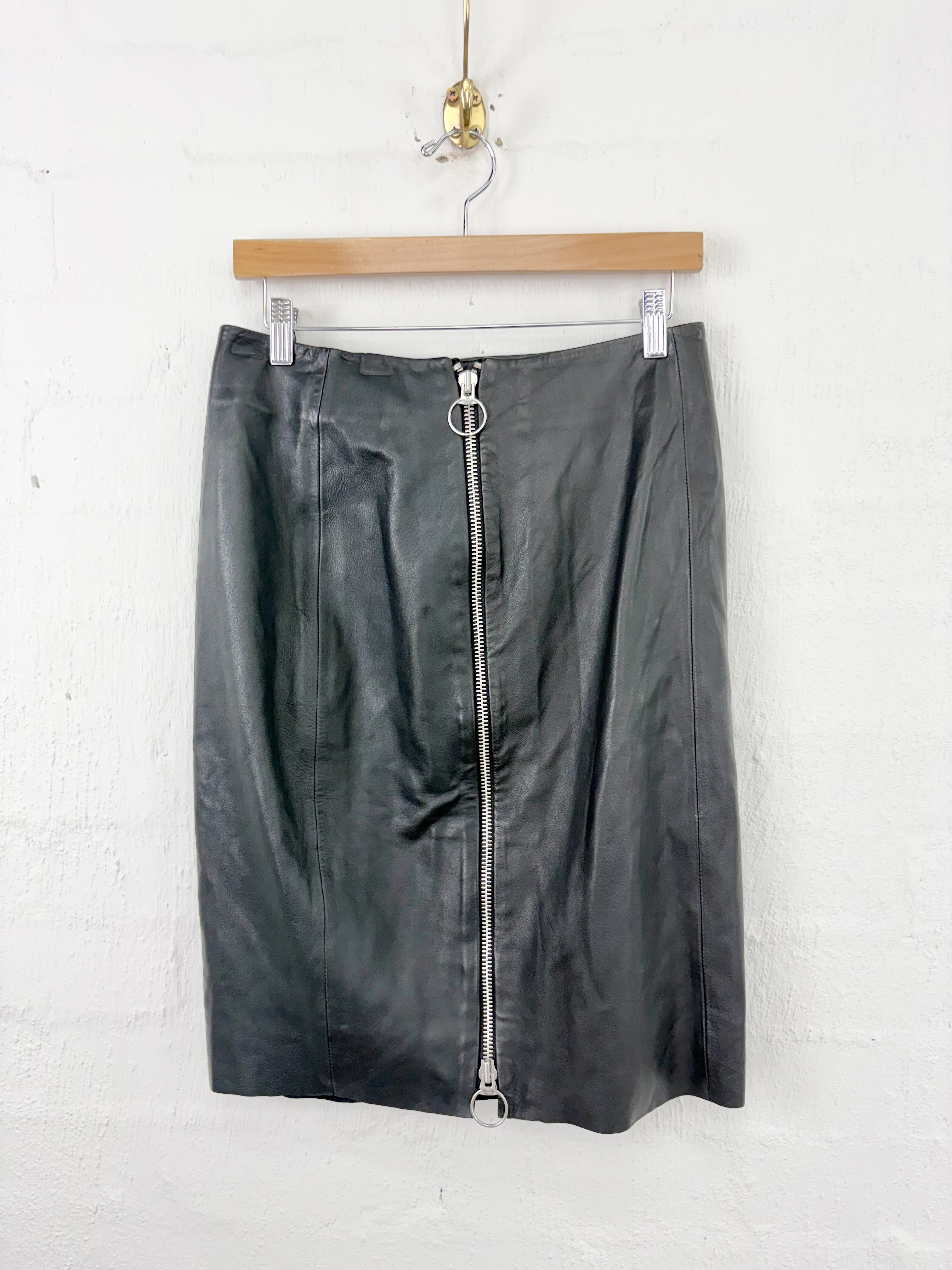 A black faux-leather pencil skirt with a prominent silver zipper down the front, hanging from a wooden hanger against a white painted brick wall.