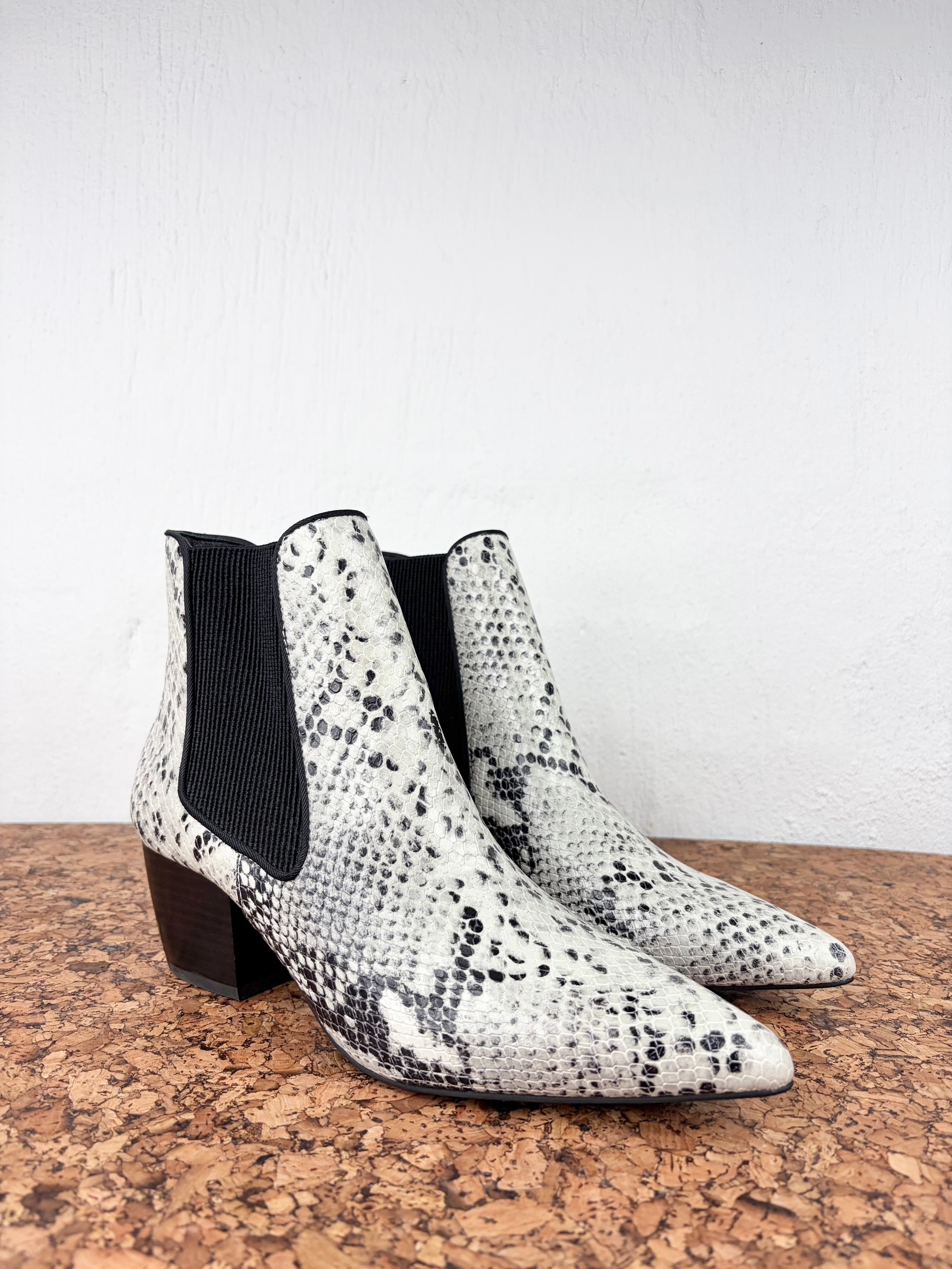 A pair of pointed-toe ankle boots with a black and white python print and black elastic side panels, resting on a cork surface against a white wall.