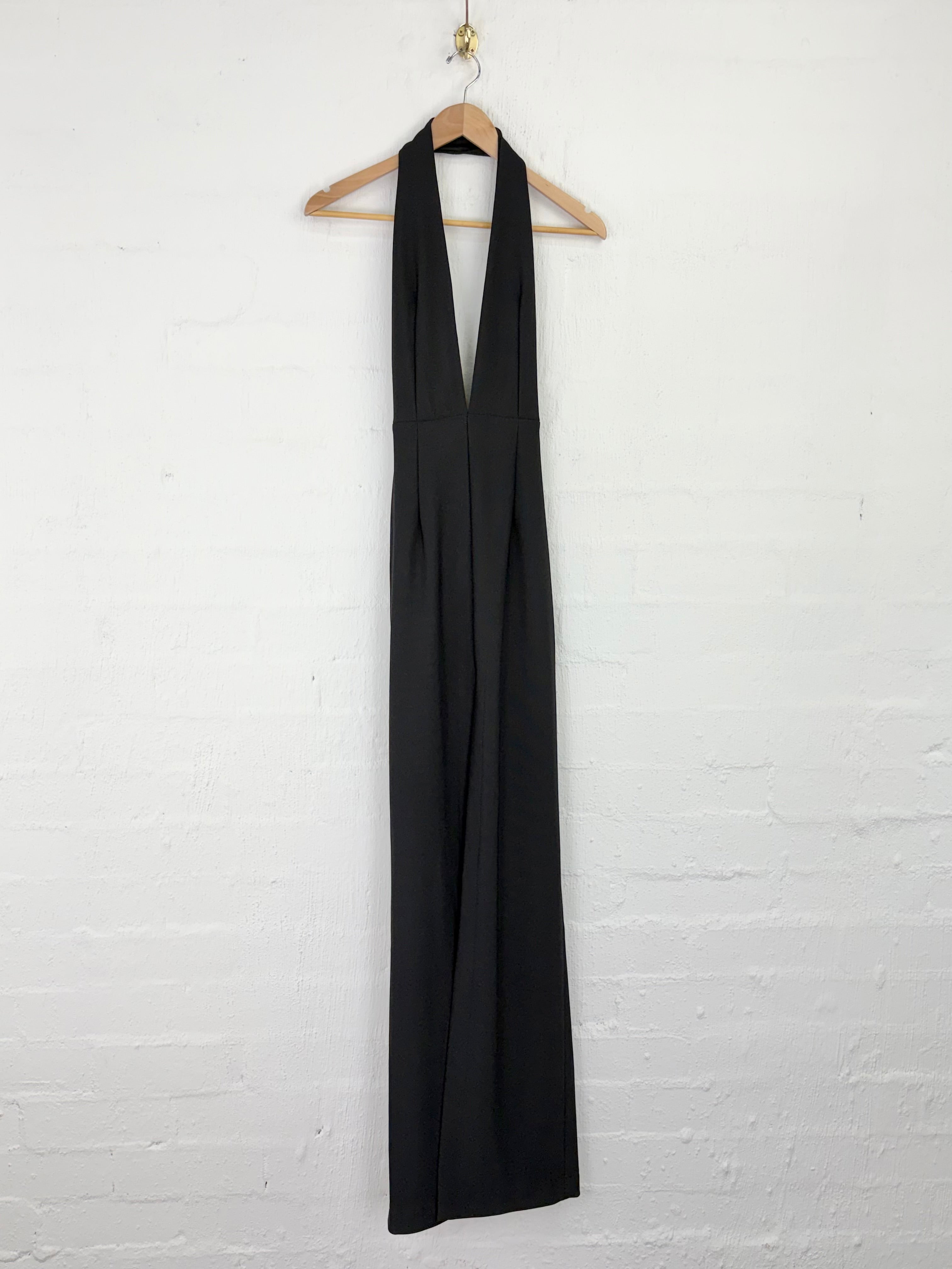 A black halter-neck maxi dress hanging on a wooden hanger against a white painted brick wall.