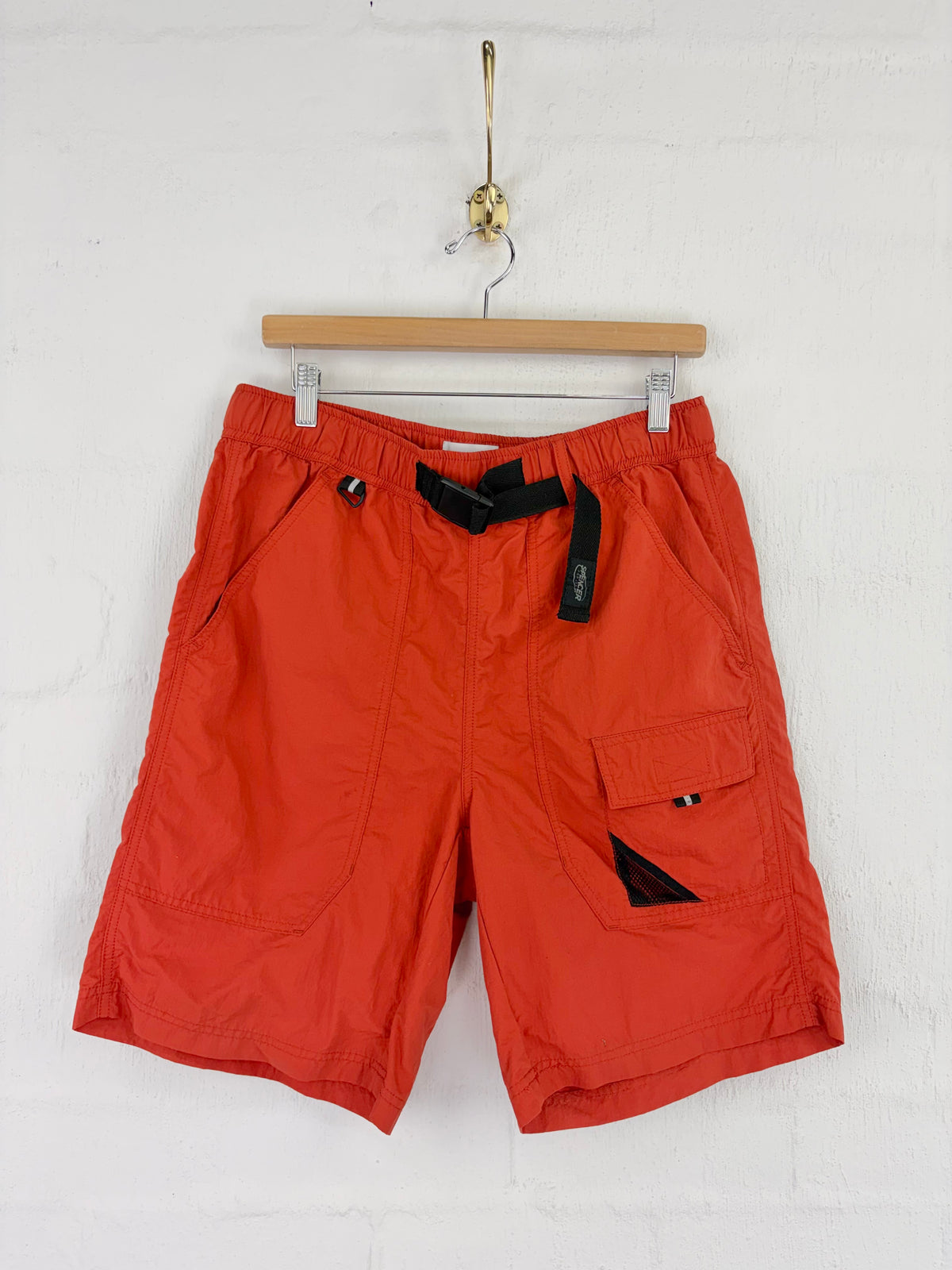 Bright orange cargo shorts with black accents and a black drawstring, hanging from a wooden hanger against a white brick wall.
