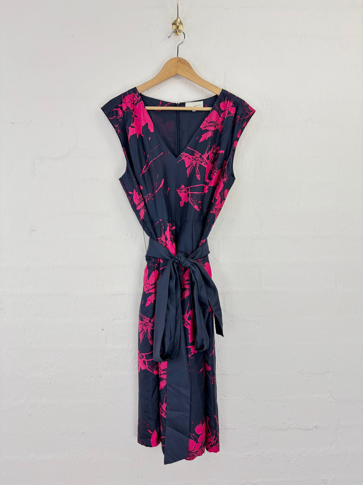 A sleeveless wrap-style dress with a navy blue base and bright pink floral print, hanging on a wooden hanger against a white painted brick wall.