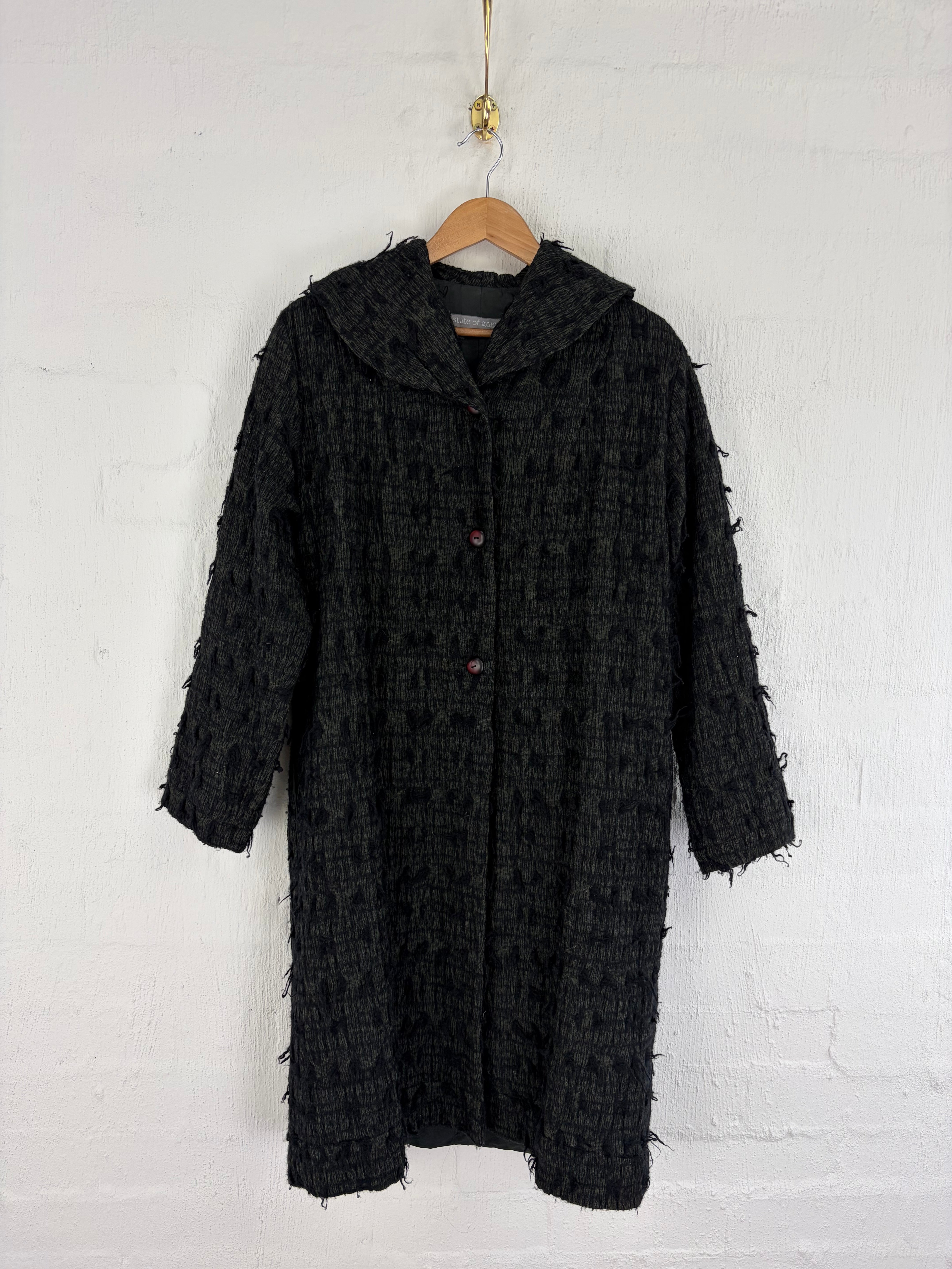 A black textured tweed coat with frayed fringe details hanging on a wooden hanger against a white painted brick wall.