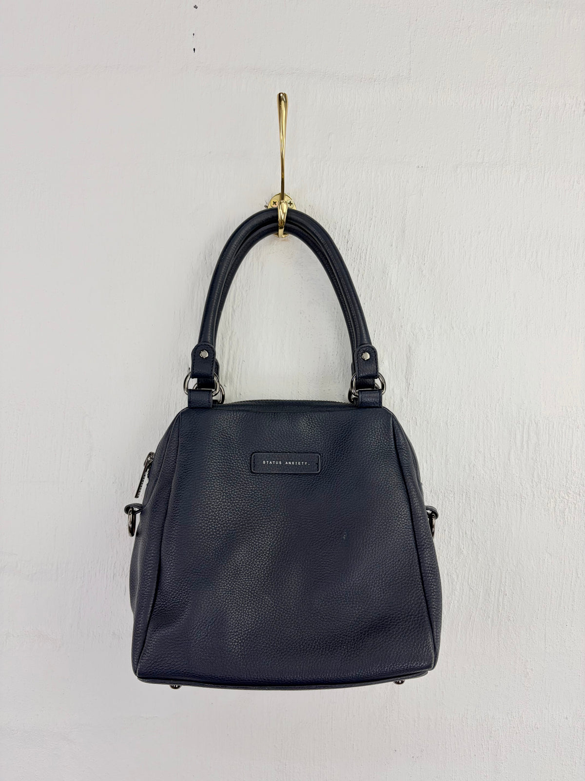 A black pebbled leather handbag with double top handles and a small silver-tone brand plaque, hanging from a gold hook against a white textured wall.