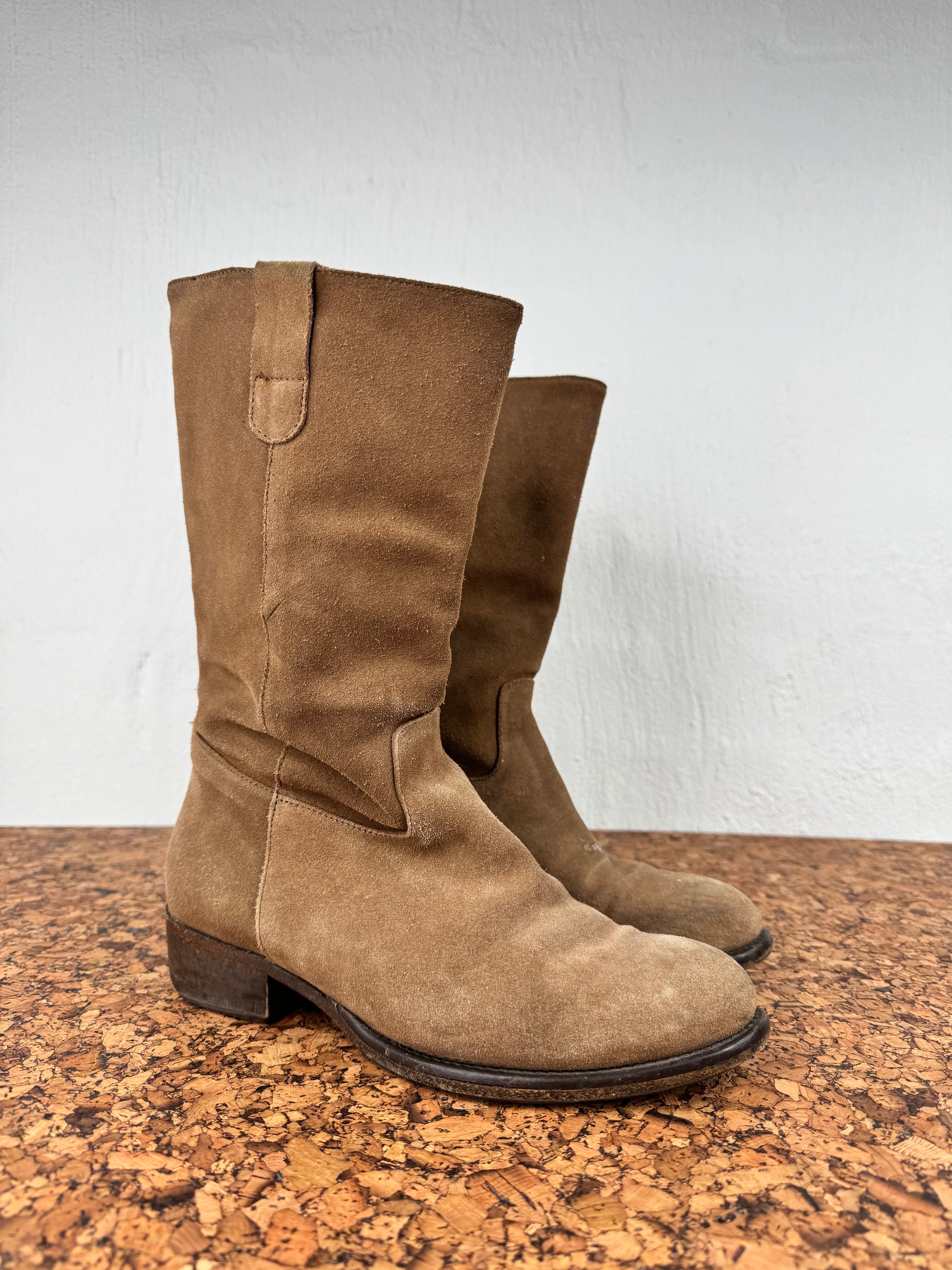 A pair of mid-calf brown suede boots with low block heels, displayed on a cork surface against a white wall.