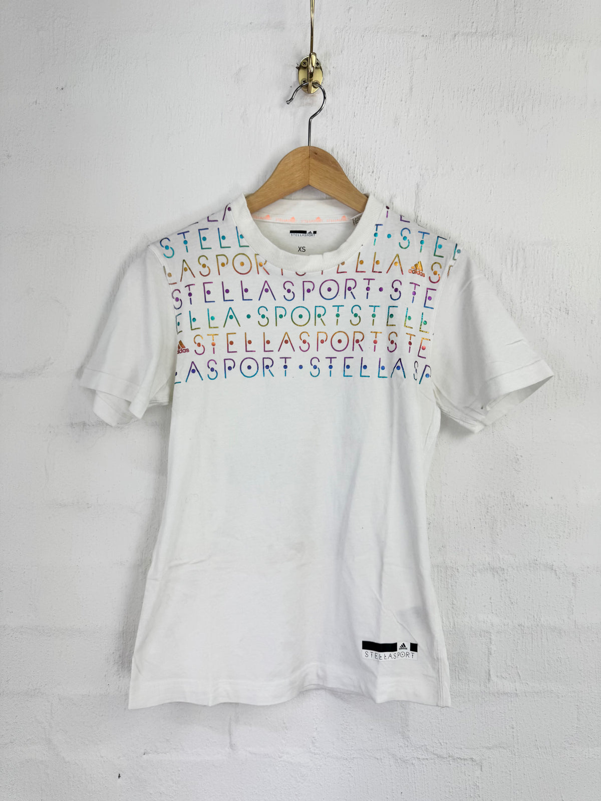 A white short-sleeve t-shirt hanging on a wooden hanger against a white painted brick wall. The shirt features a colorful graphic print across the chest with the word 'STELLA' repeated in various colors (blue, green, orange, purple, red).